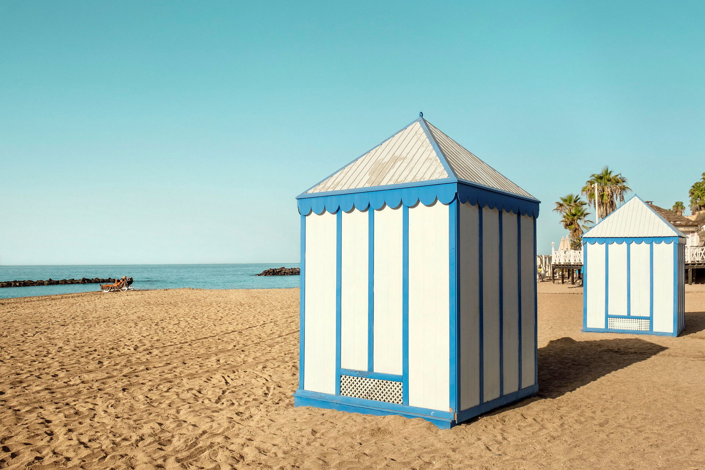 Två strandpaviljonger med blå och vita väggar står på en sandstrand vid havet. (AI-genererad text)