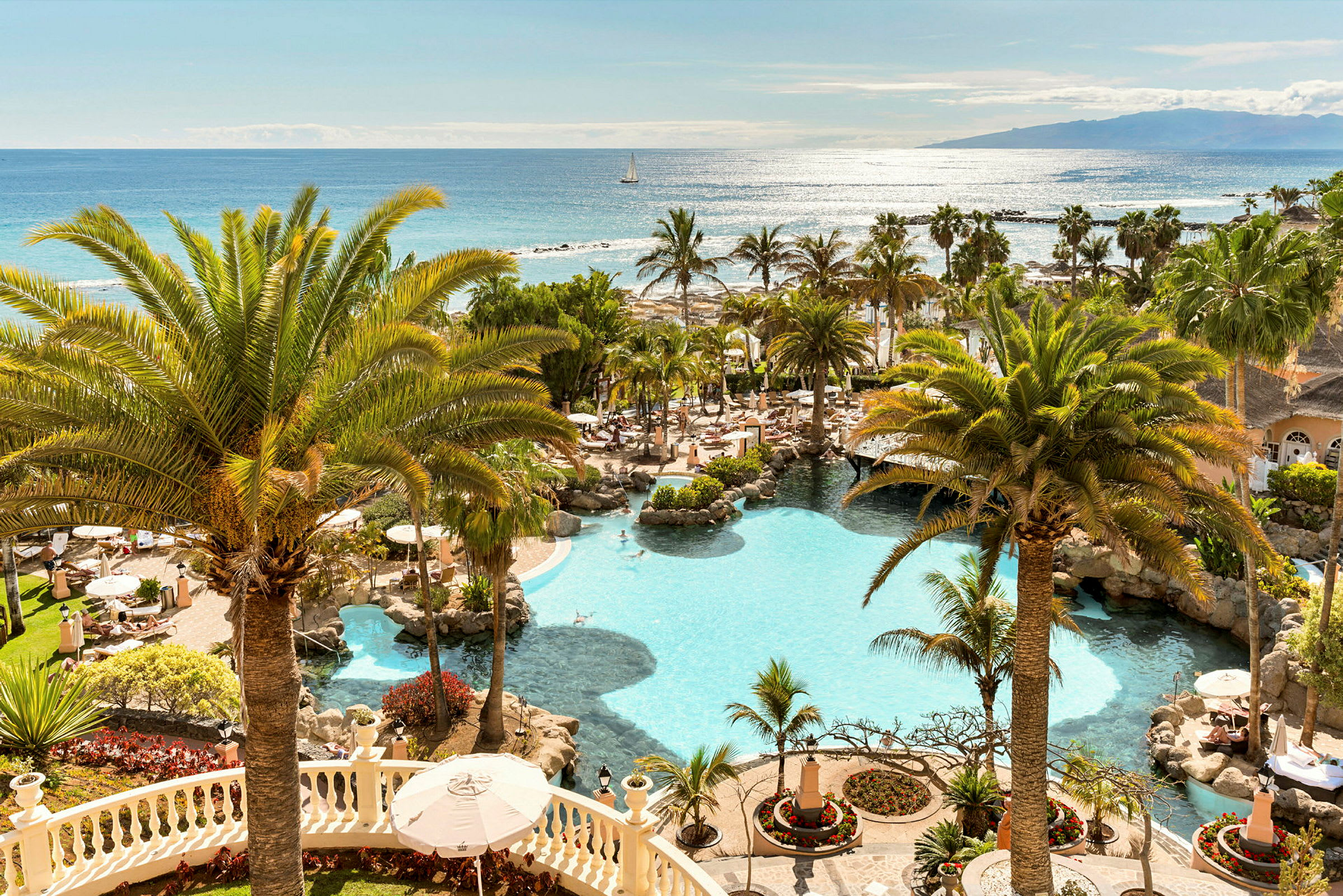 En utsikt över en hotellpool omgiven av palmer och träd, med havet i bakgrunden. (AI-genererad text)