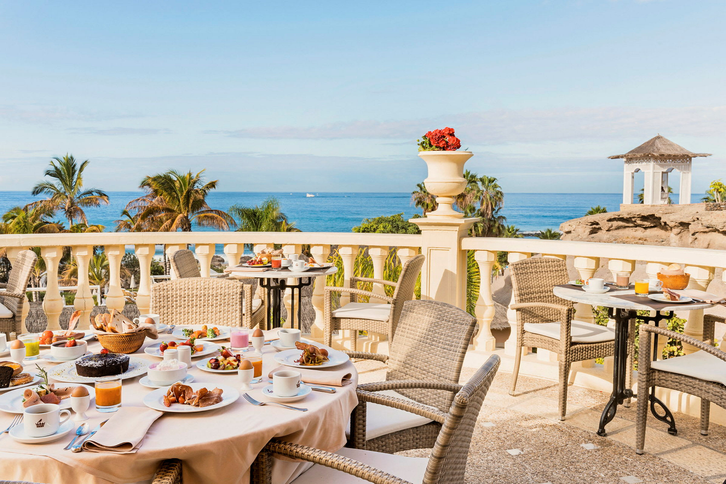 Frukostbord med olika maträtter på en terrass med utsikt över palmer och havet. (AI-genererad text)