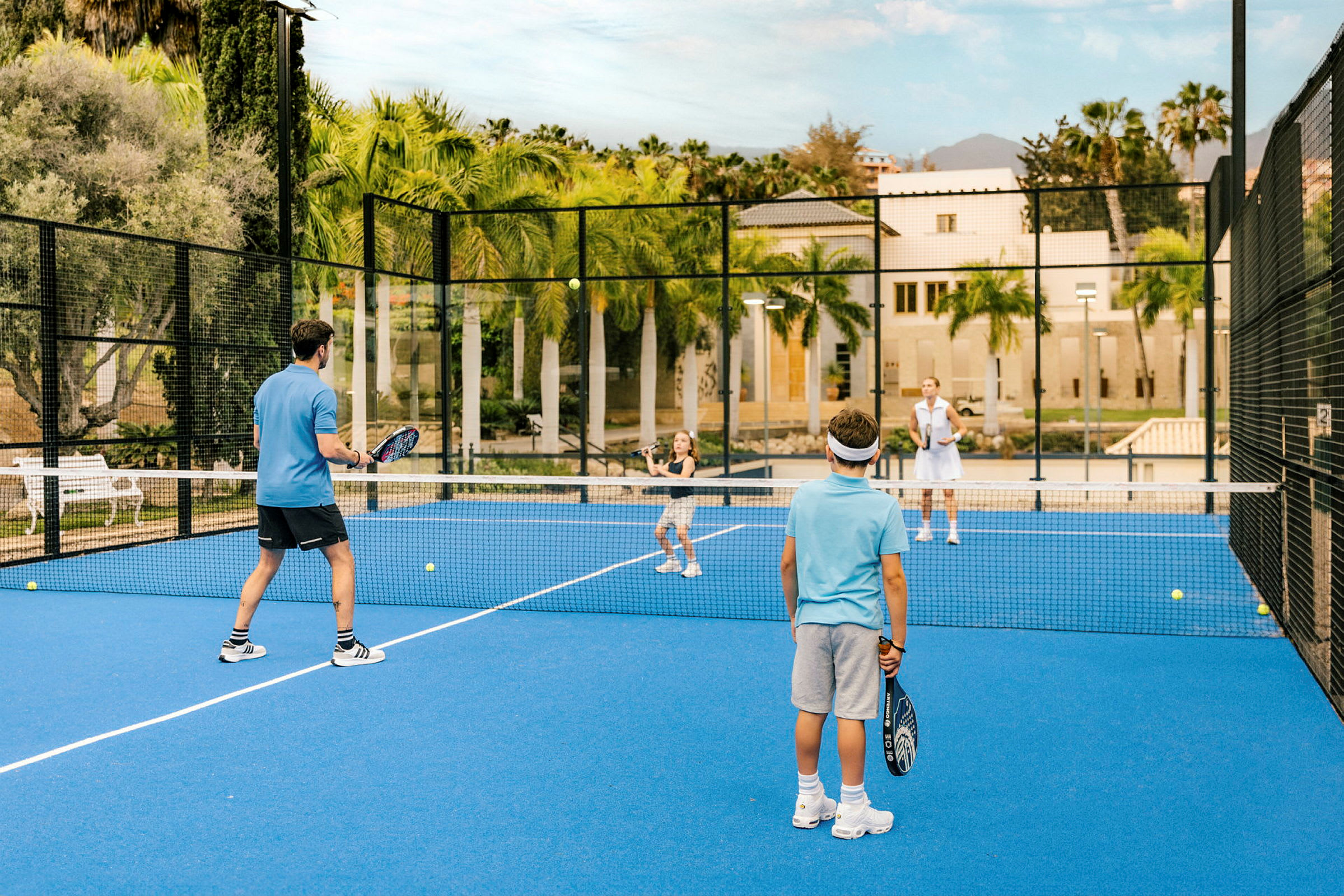 Fyra personer spelar padel på en blå bana omgiven av nät och palmer. (AI-genererad text)
