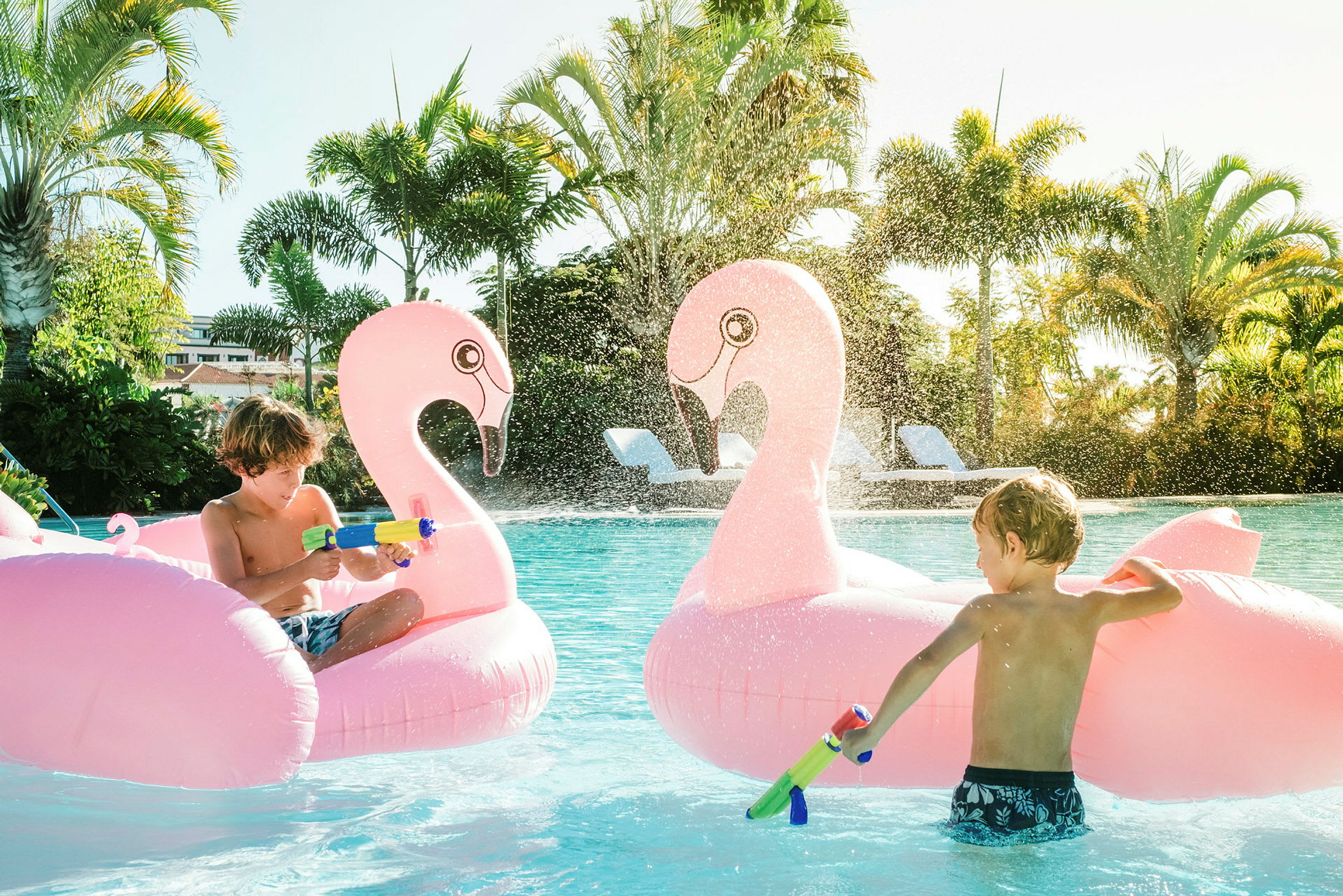 Två uppblåsbara flamingo-figurer i en pool med vattenstrålar och omgivande palmer. (AI-genererad text)