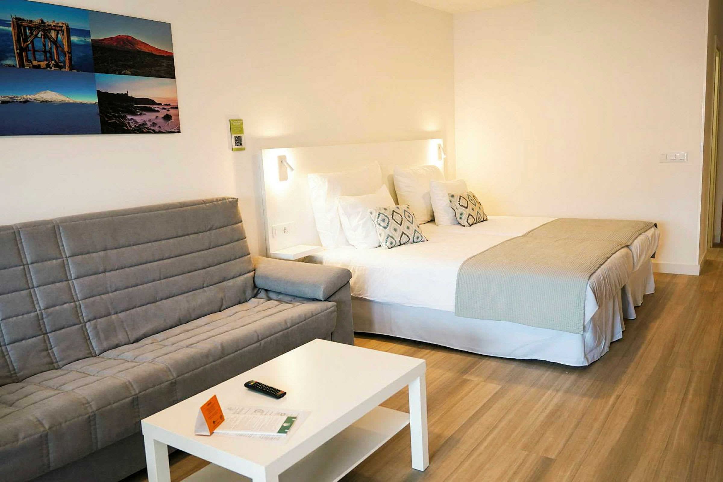 Hotellrum med en dubbelsäng, en grå soffa, ett vitt bord och bilder på väggen. (AI-genererad text)