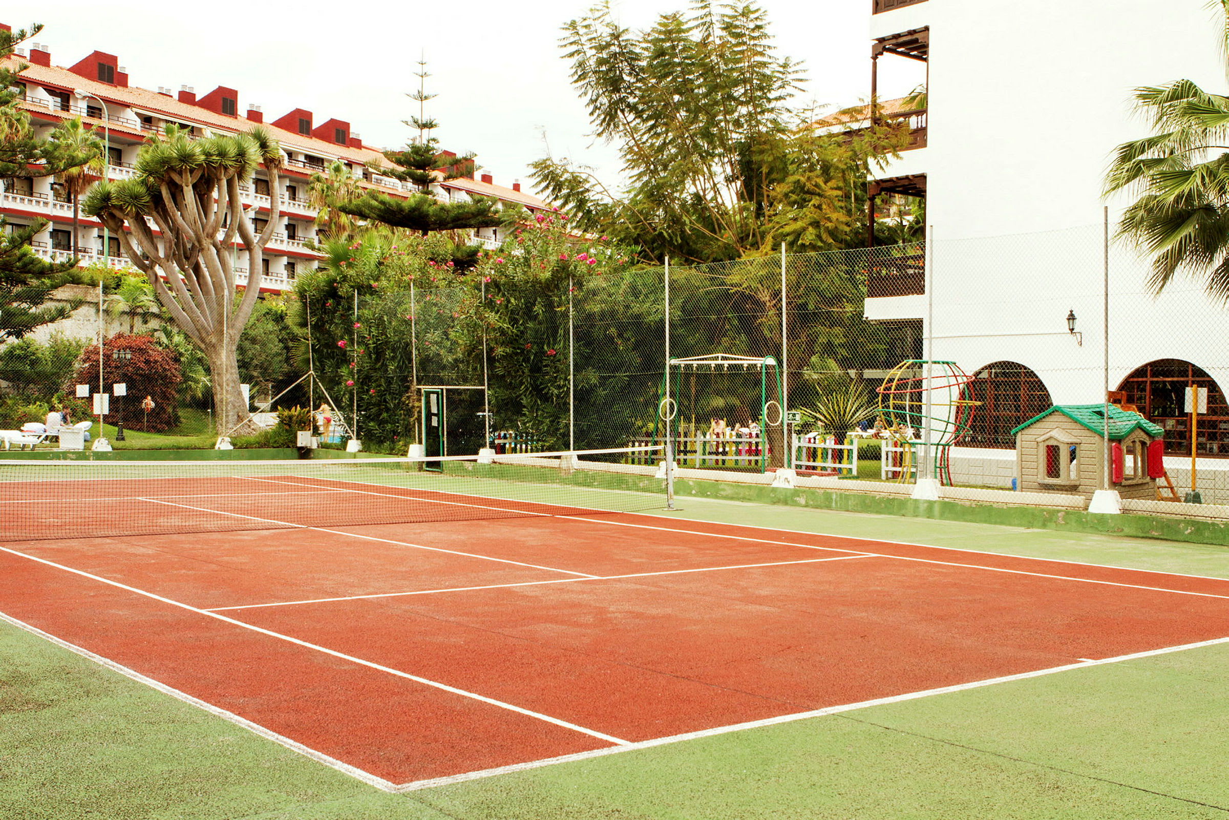 En tennisbana omgiven av grönska och byggnader. (AI-genererad text)