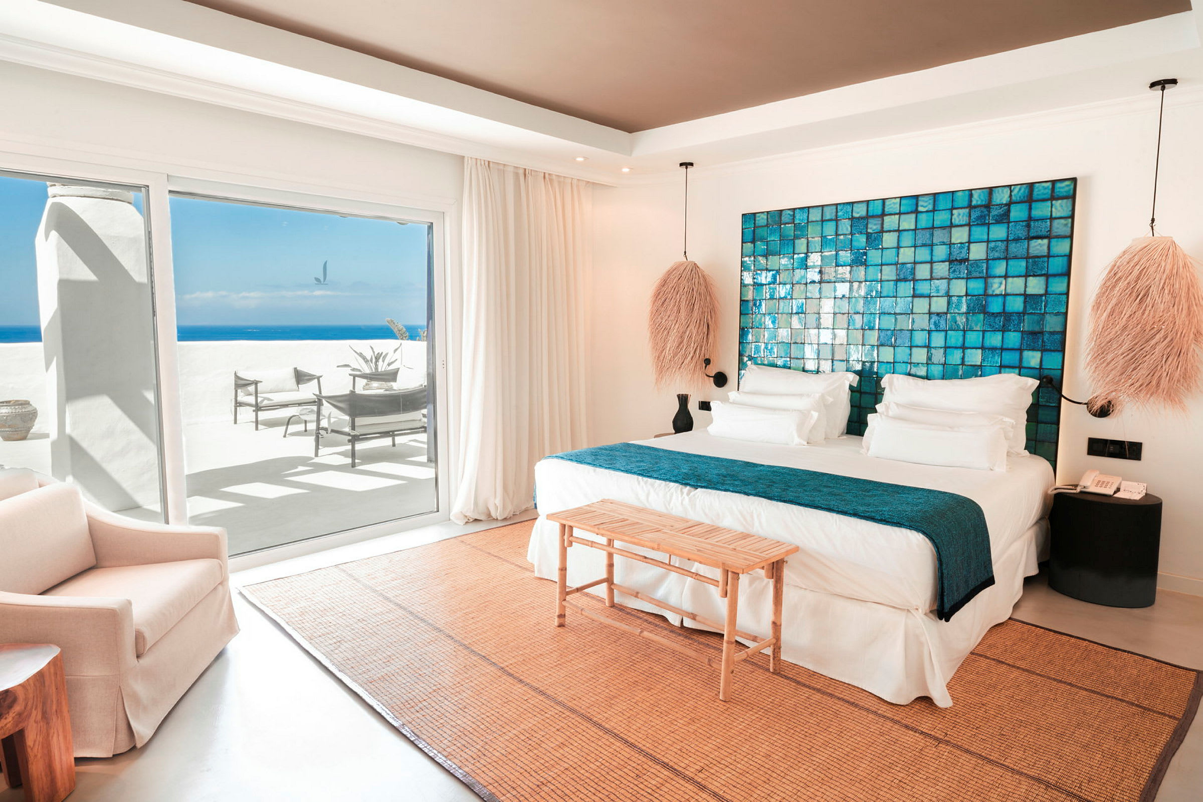 Ett hotellrum med en dubbelsäng, blått dekorativt huvudgavel, sittmöbler och utgång mot en terrass med havsutsikt. (AI-genererad text)