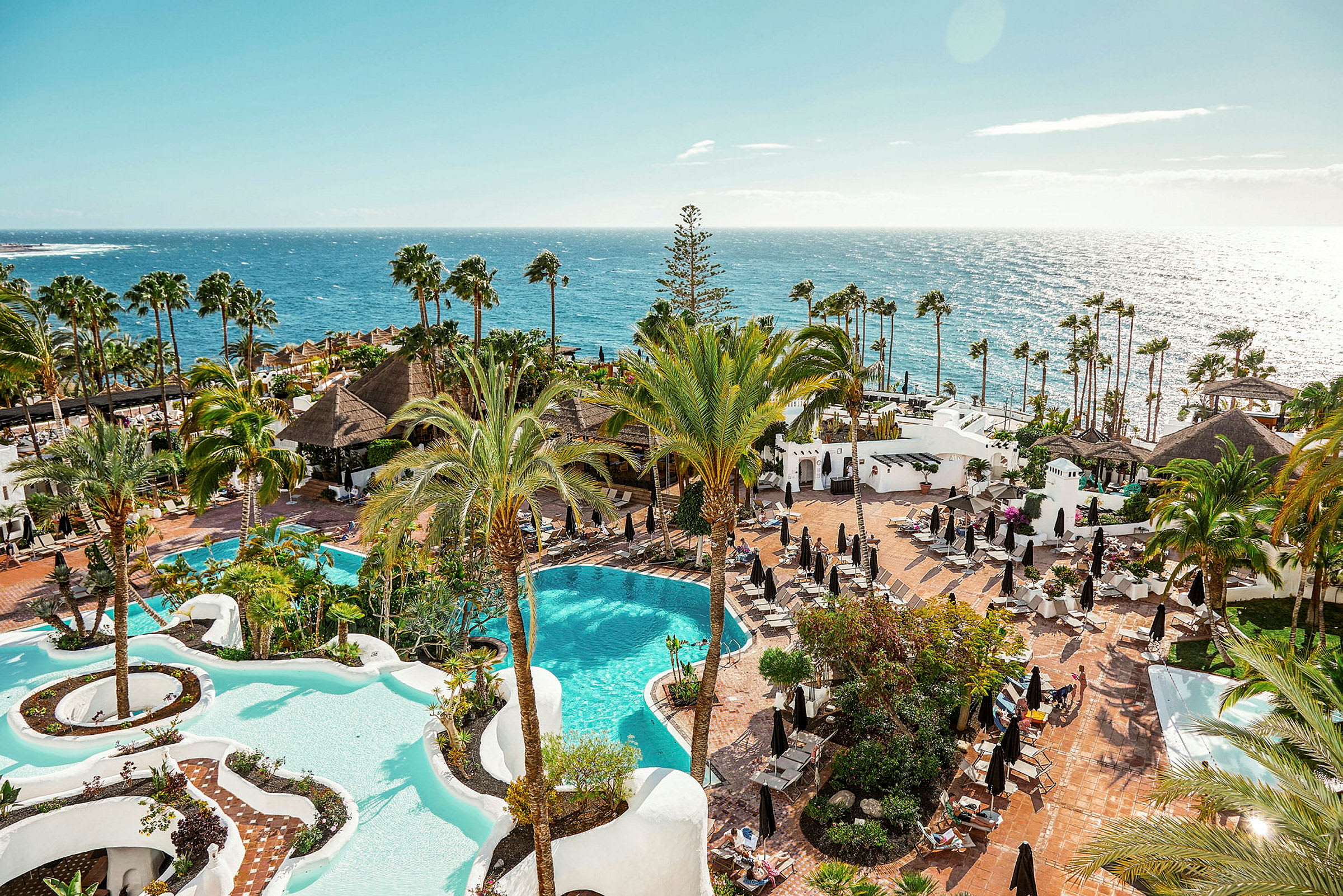 Vy över ett hotellområde med två pooler, palmer och solsängar vid havet. (AI-genererad text)