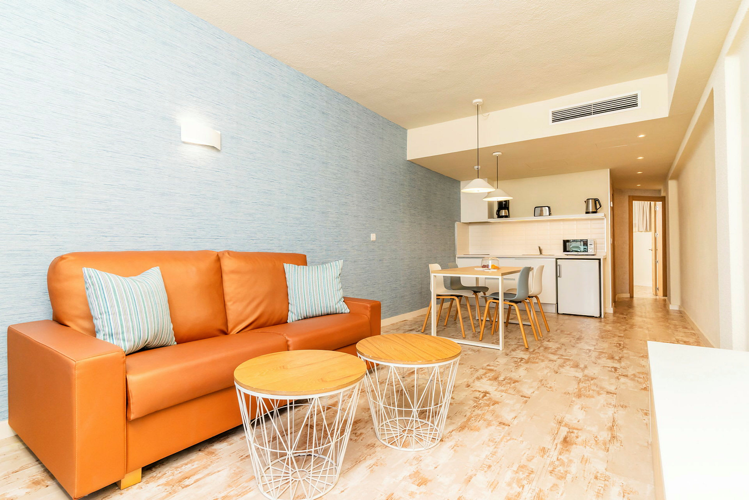 Bild på ett vardagsrum med en orange soffa, två små soffbord, och ett matbord med stolar i en öppen planlösning som inkluderar ett kök. (AI-genererad text)