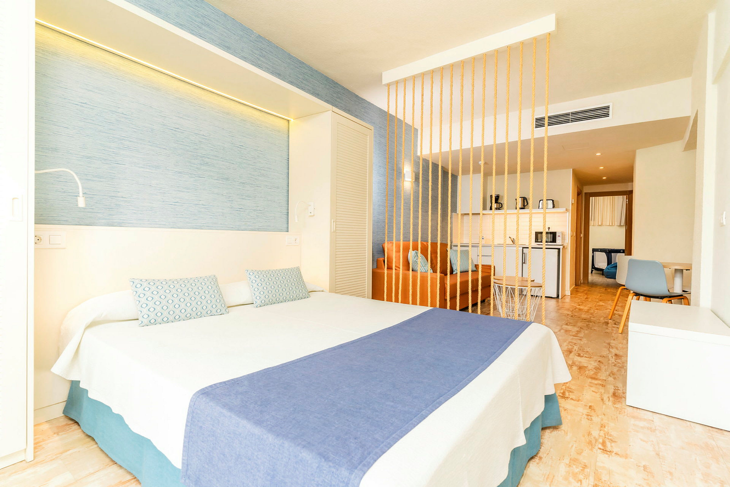 Hotellrum med en dubbelsäng, blått överkast och dekorativa kuddar, samt en orange soffa bakom en dekorativ avskärmningsvägg. (AI-genererad text)