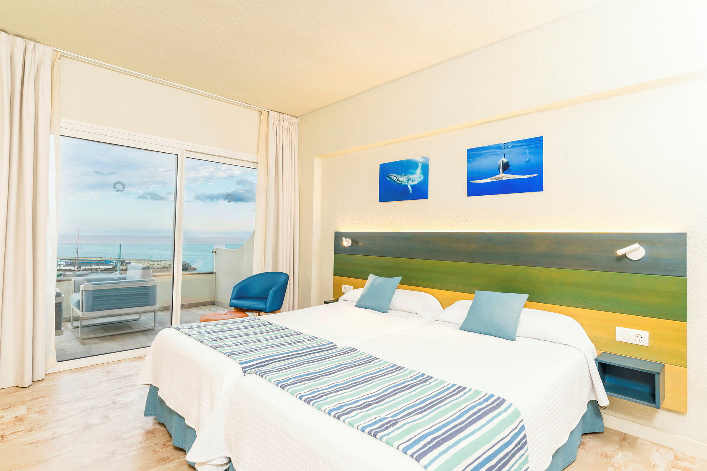 Hotelrum med två enkelsängar, en grön och blå sänggavel, samt en balkong med utsikt över havet. (AI-genererad text)