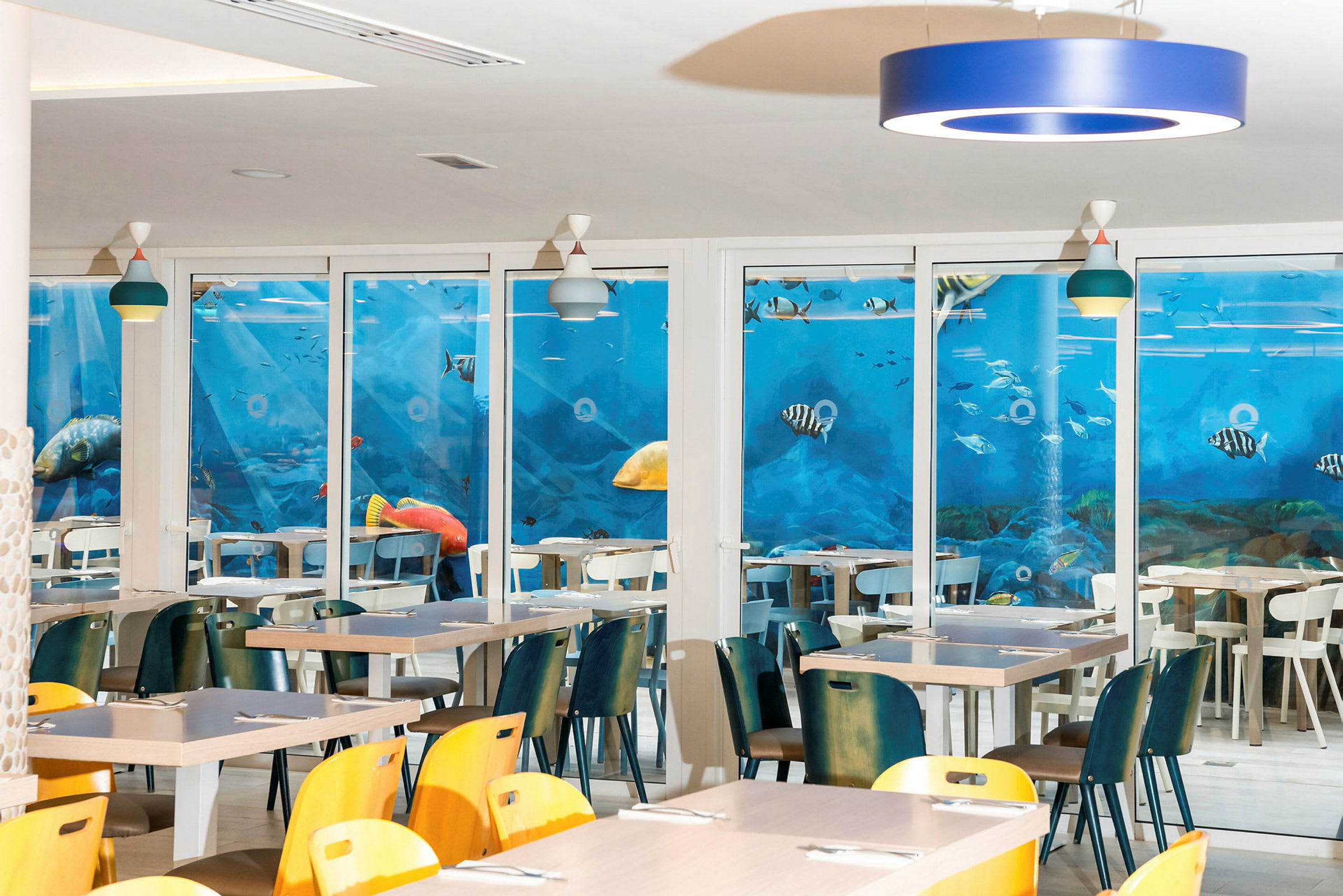 Restaurang med bord och stolar, med en vägg täckt av ett blått akvarie-motiv. (AI-genererad text)