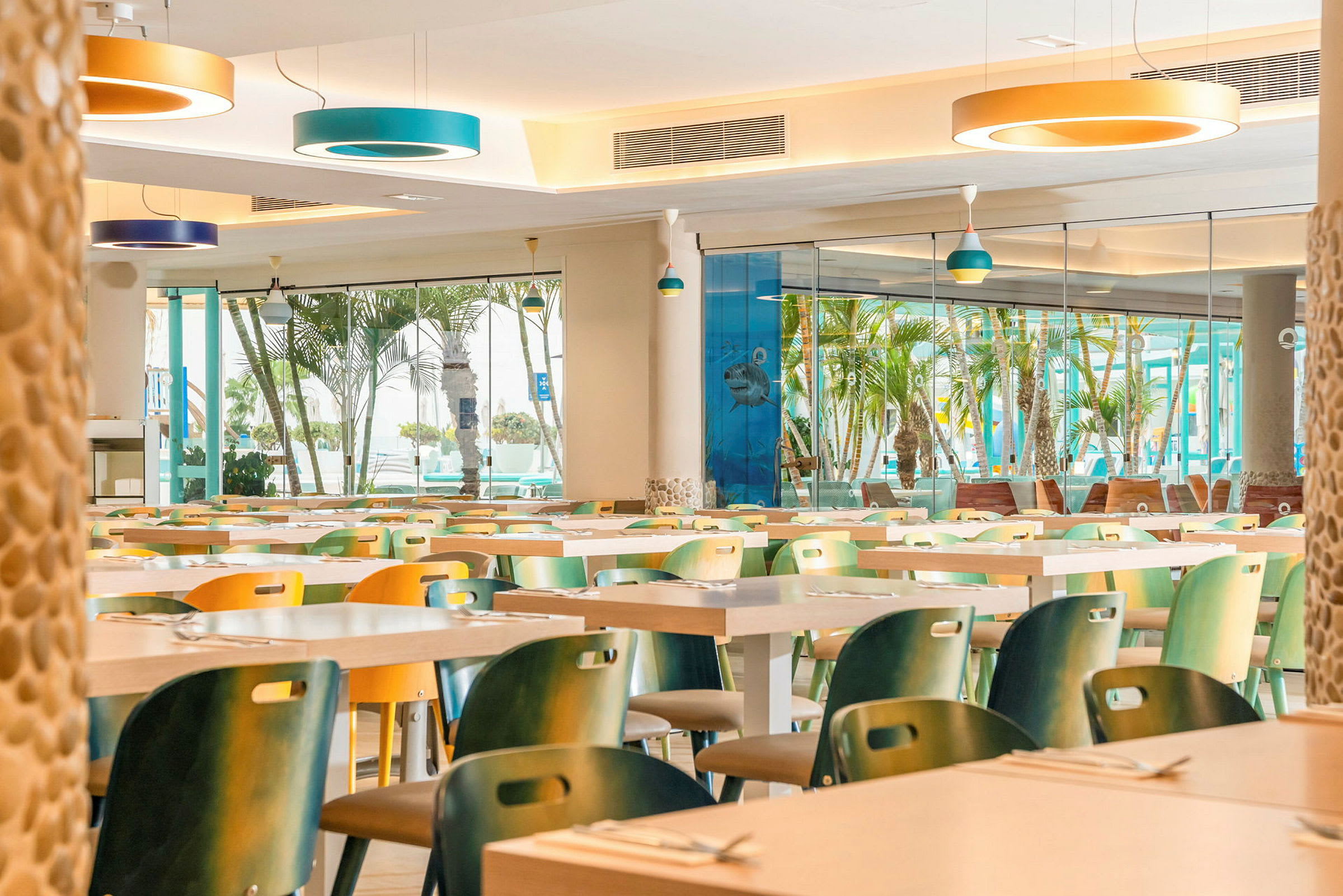 Restaurang med färgglada stolar och stora fönster som vetter mot en utsikt med palmer. (AI-genererad text)