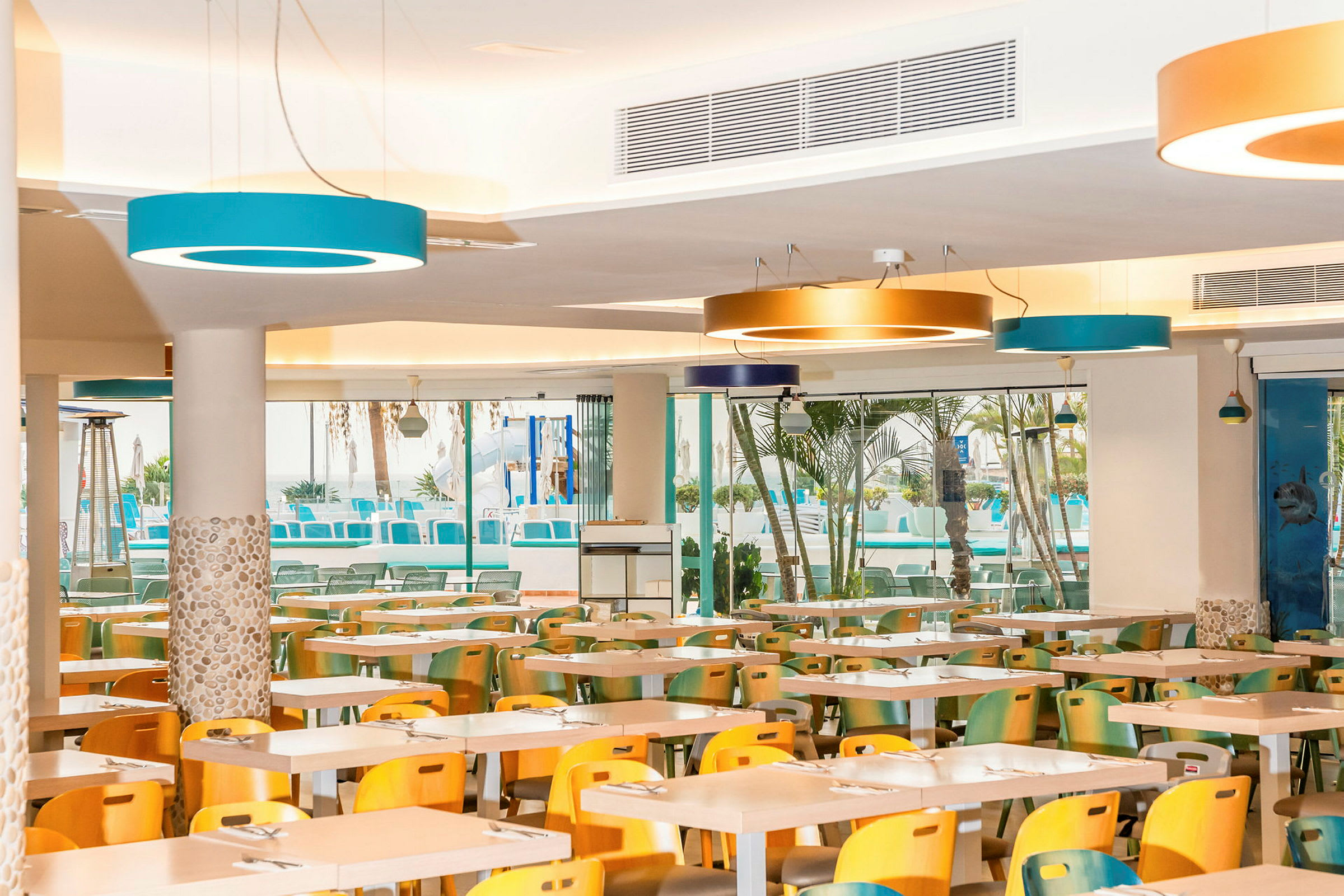 En restaurang med färgglada stolar och bord, samt stora fönster mot en utomhuspool. (AI-genererad text)
