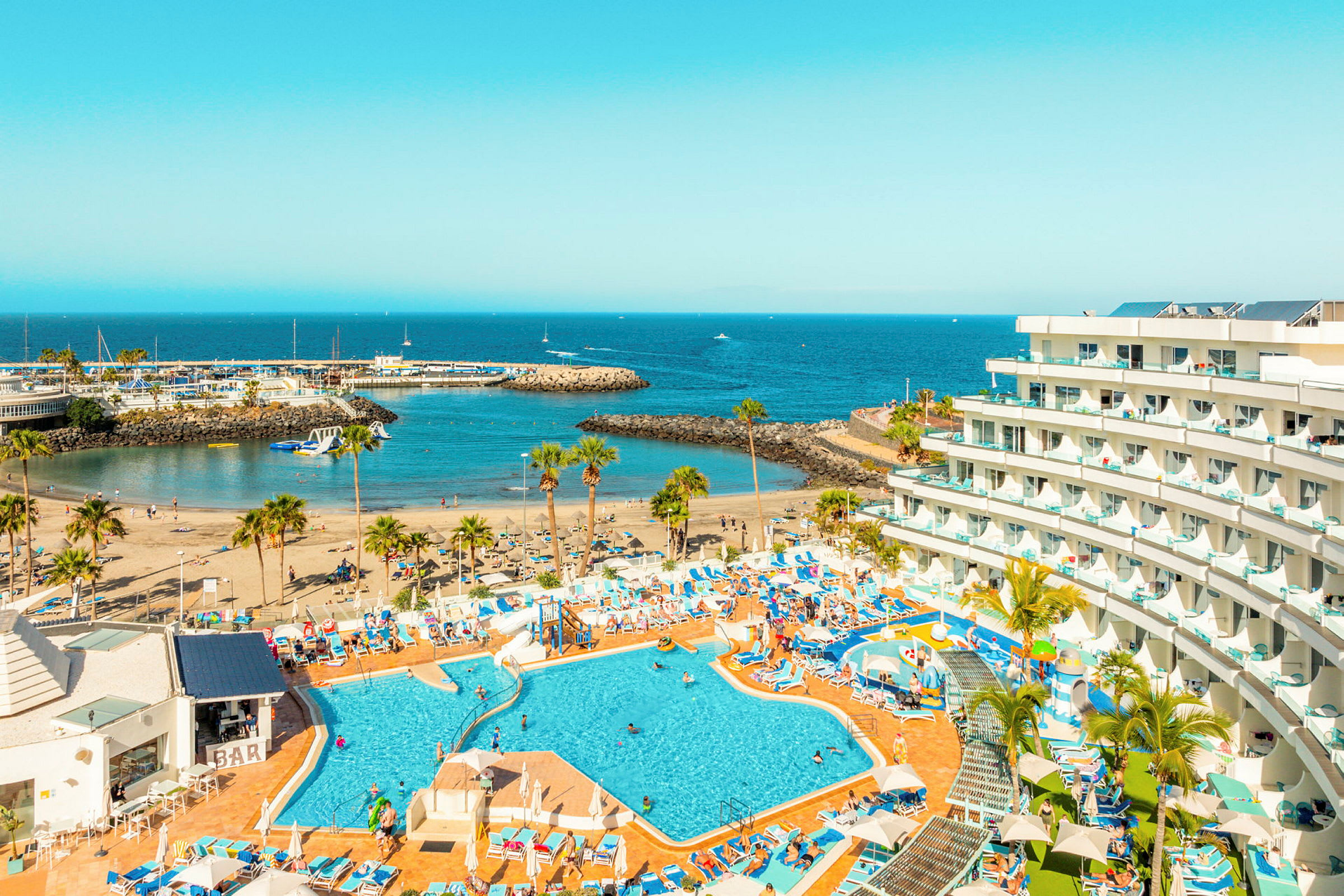 Bild av ett hotell med poolområde, strand och hamninlopp vid havet. (AI-genererad text)