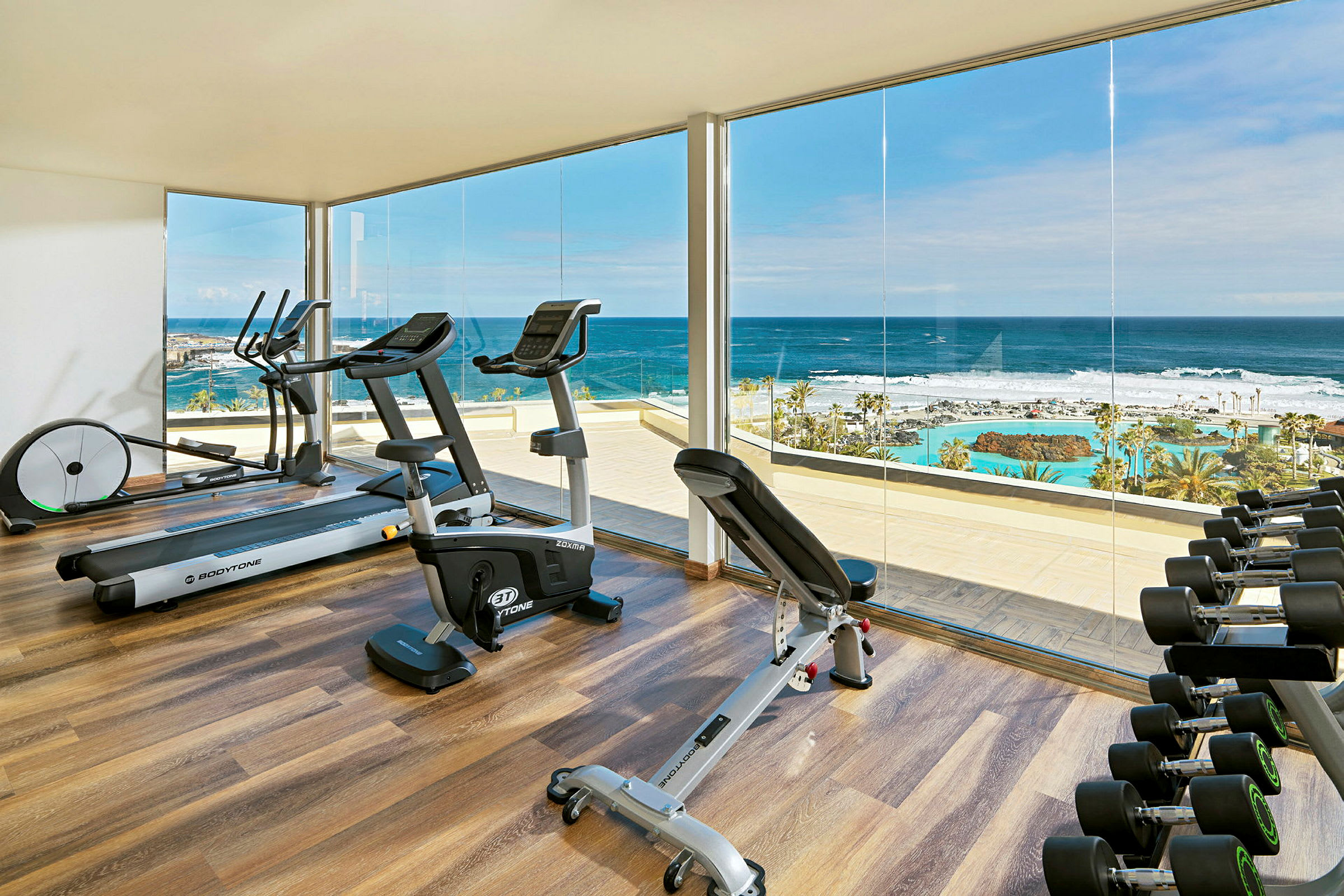Ett gym med träningsutrustning, stora fönster och utsikt över havet och ett poolområde. (AI-genererad text)