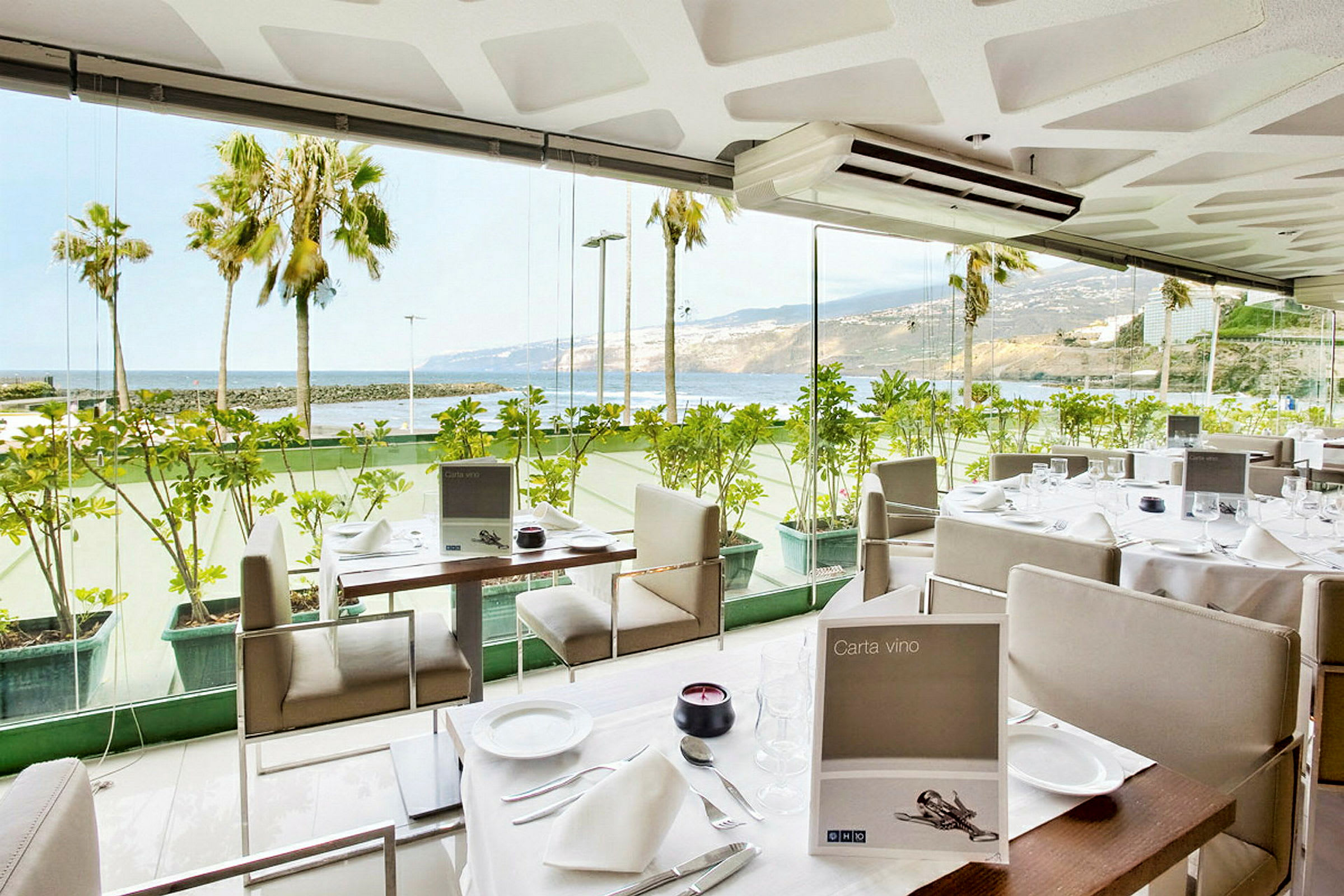 En restaurang med glasväggar och utsikt mot havet och palmer. (AI-genererad text)