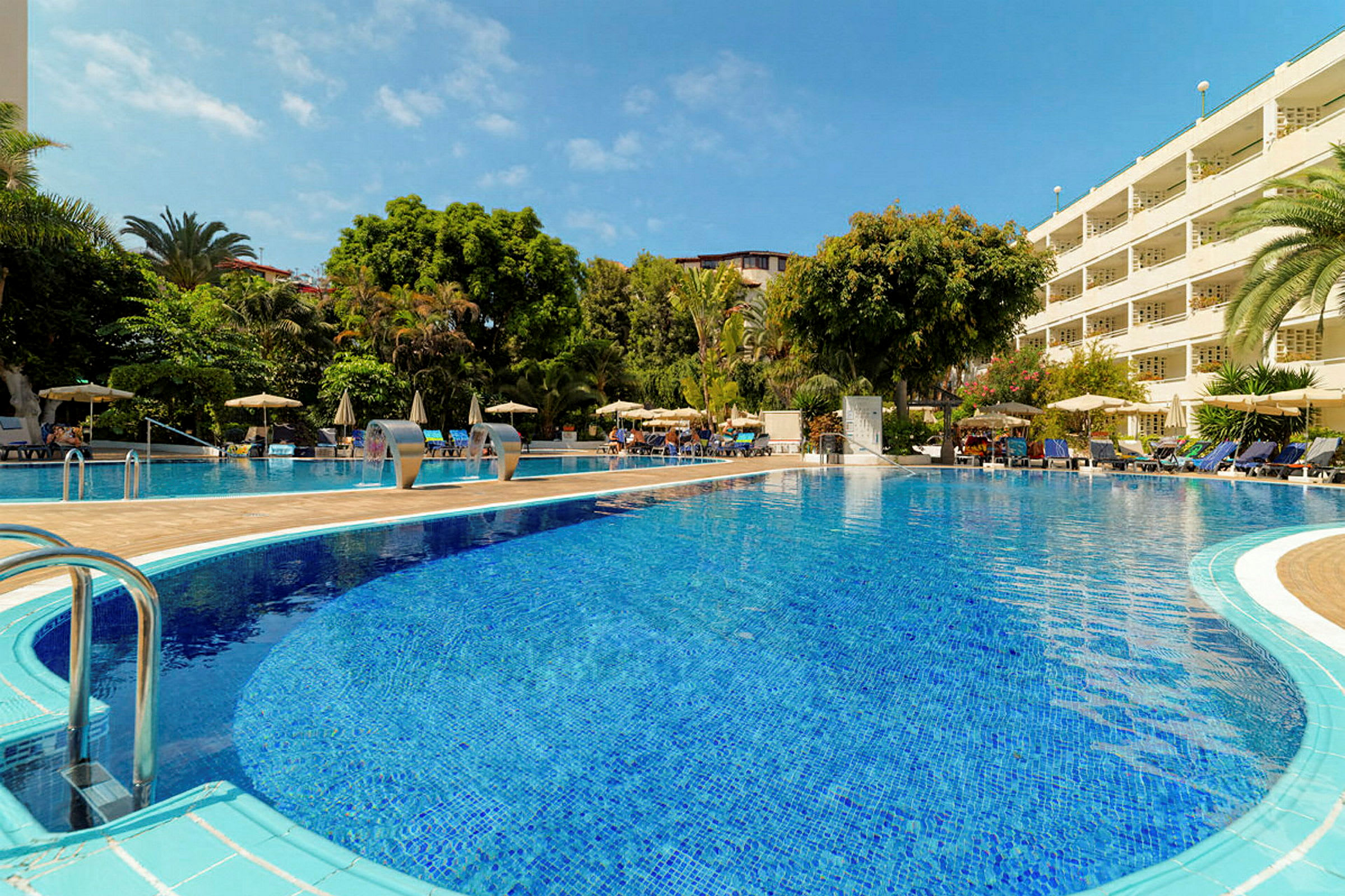 En solig hotellpool omgiven av grönska och solstolar. (AI-genererad text)
