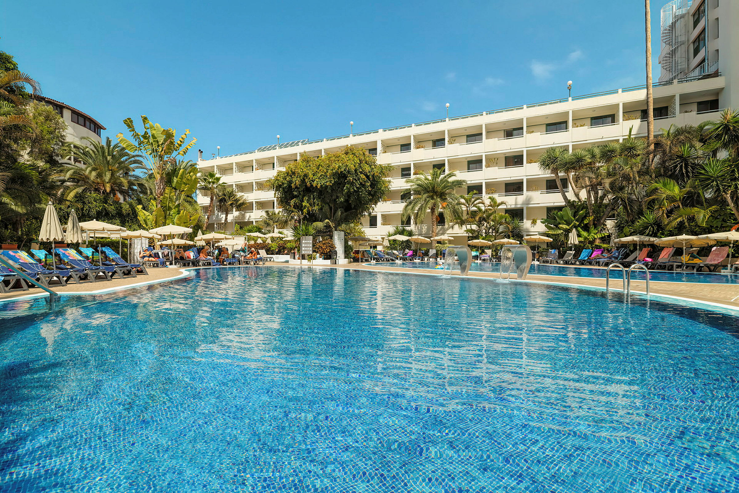En pool framför ett hotell omgivet av palmer och solstolar. (AI-genererad text)