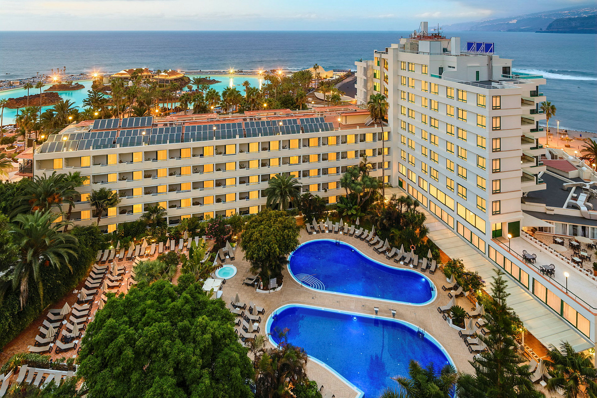 Ett hotellkomplex med pooler och grönska i förgrunden samt en kustlinje och hav i bakgrunden. (AI-genererad text)