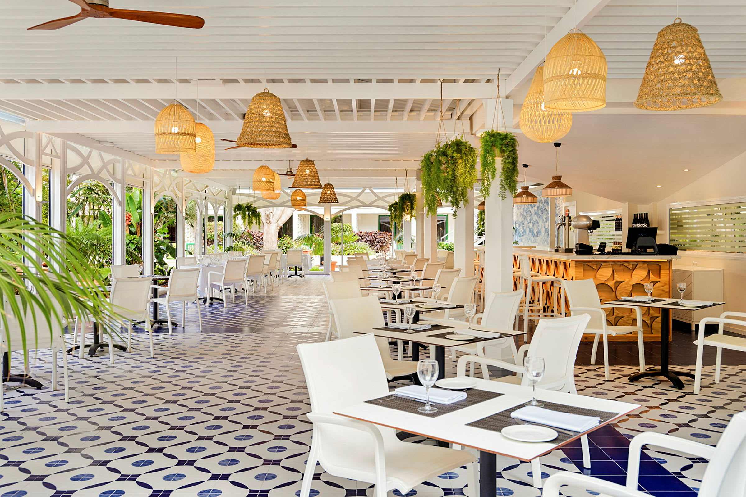 Bild av en restaurang med vita möbler, dekorativt kaklat golv och taklampor i rotting. Gröna växter hänger från taket. (AI-genererad text)