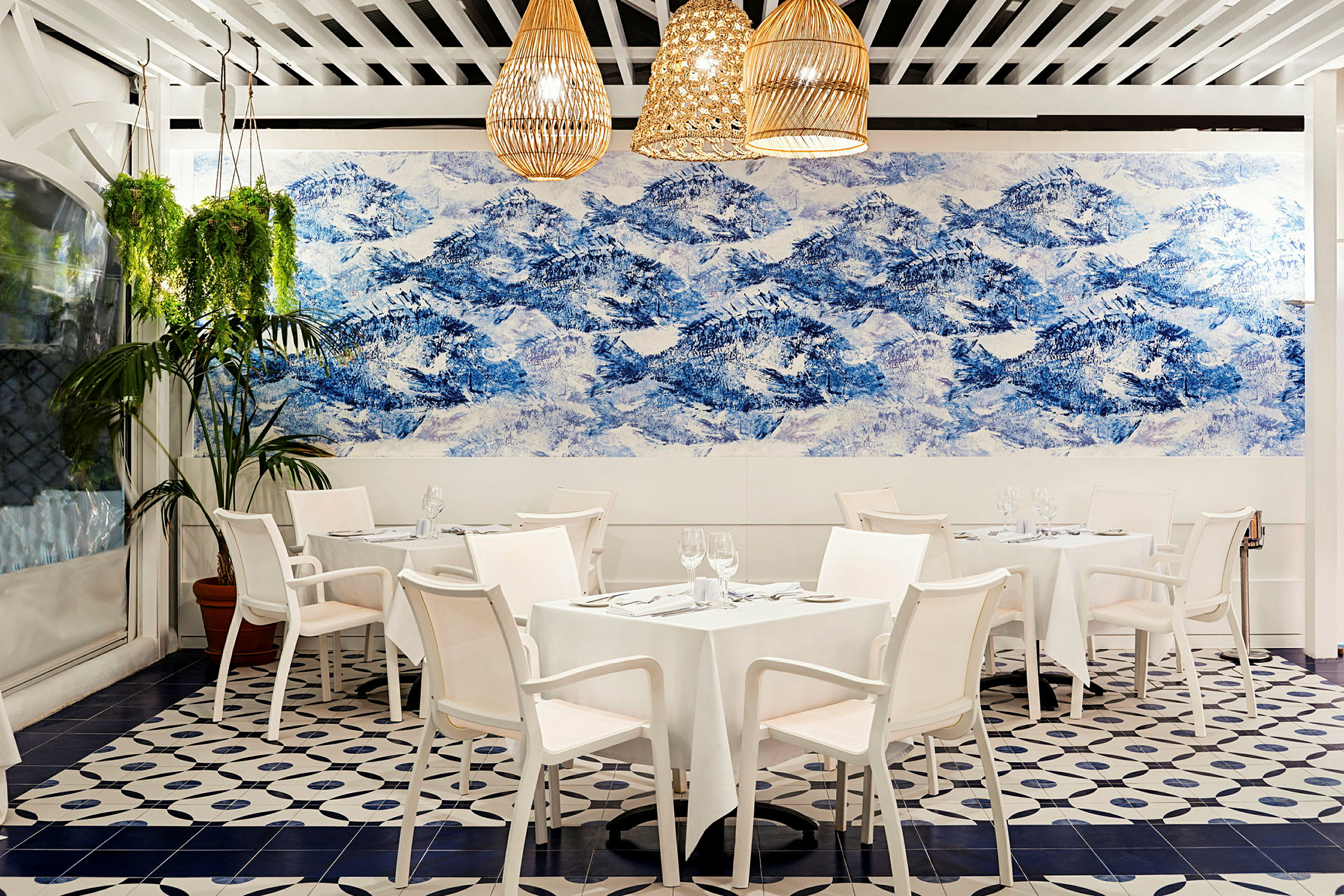 En inomhus restaurang med vita bord och stolar, dekorerad med en stor väggmålning i blått och vitt som föreställer berg. Hängande växter pryder lokalen. (AI-genererad text)