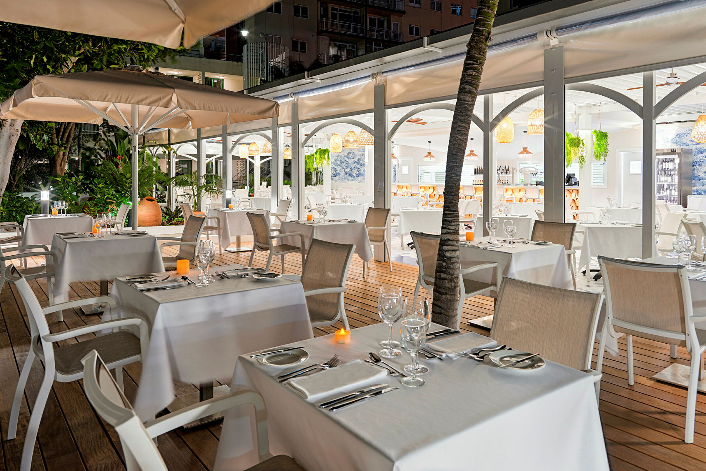 En utomhusrestaurang med dukade bord och stolar, omgiven av grönska och belyst av mjukt ljus. (AI-genererad text)