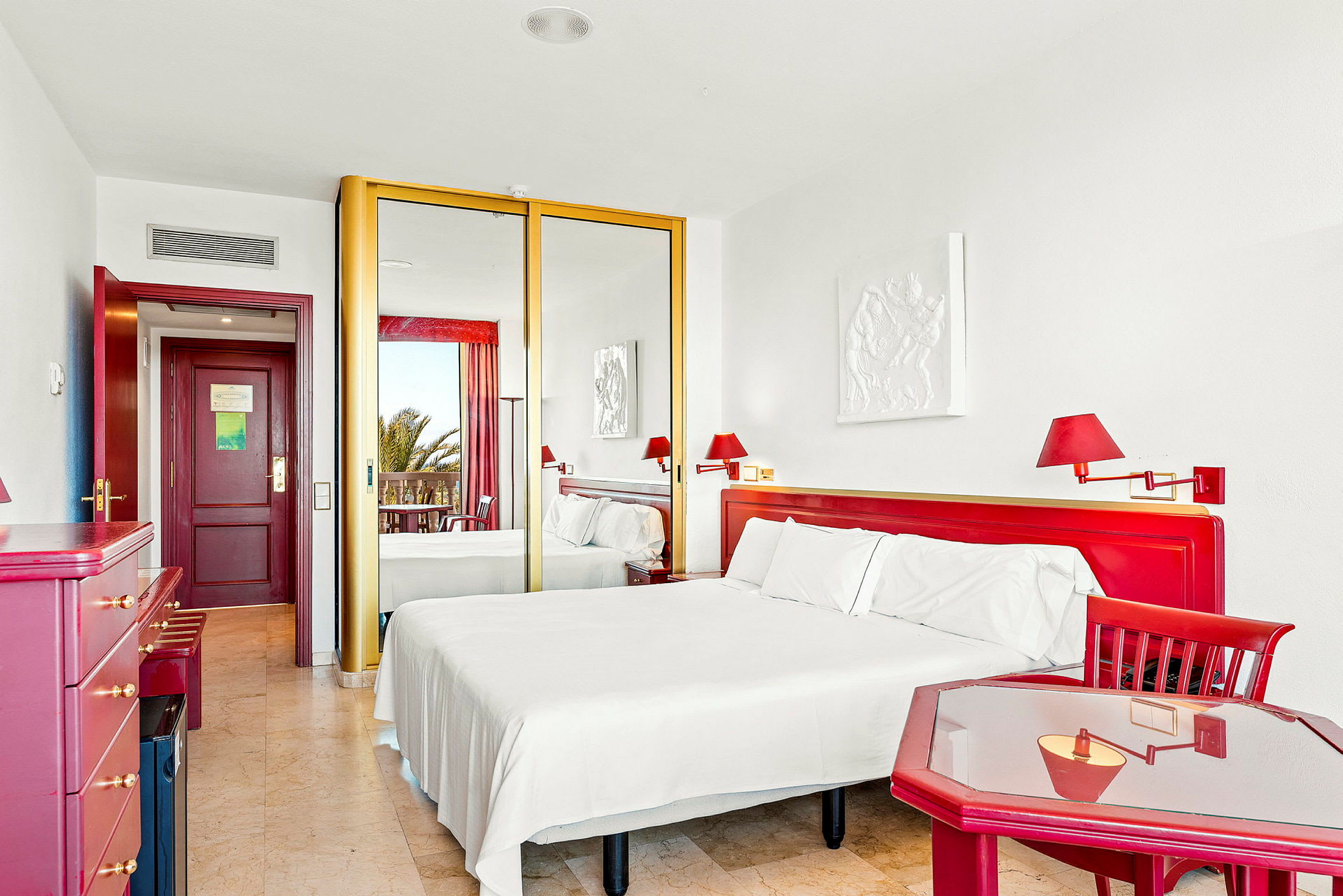 Ett hotellrum med en dubbel säng, röda möbler och speglar som garderobsdörrar. Ett fönster eller balkong syns i bakgrunden. (AI-genererad text)