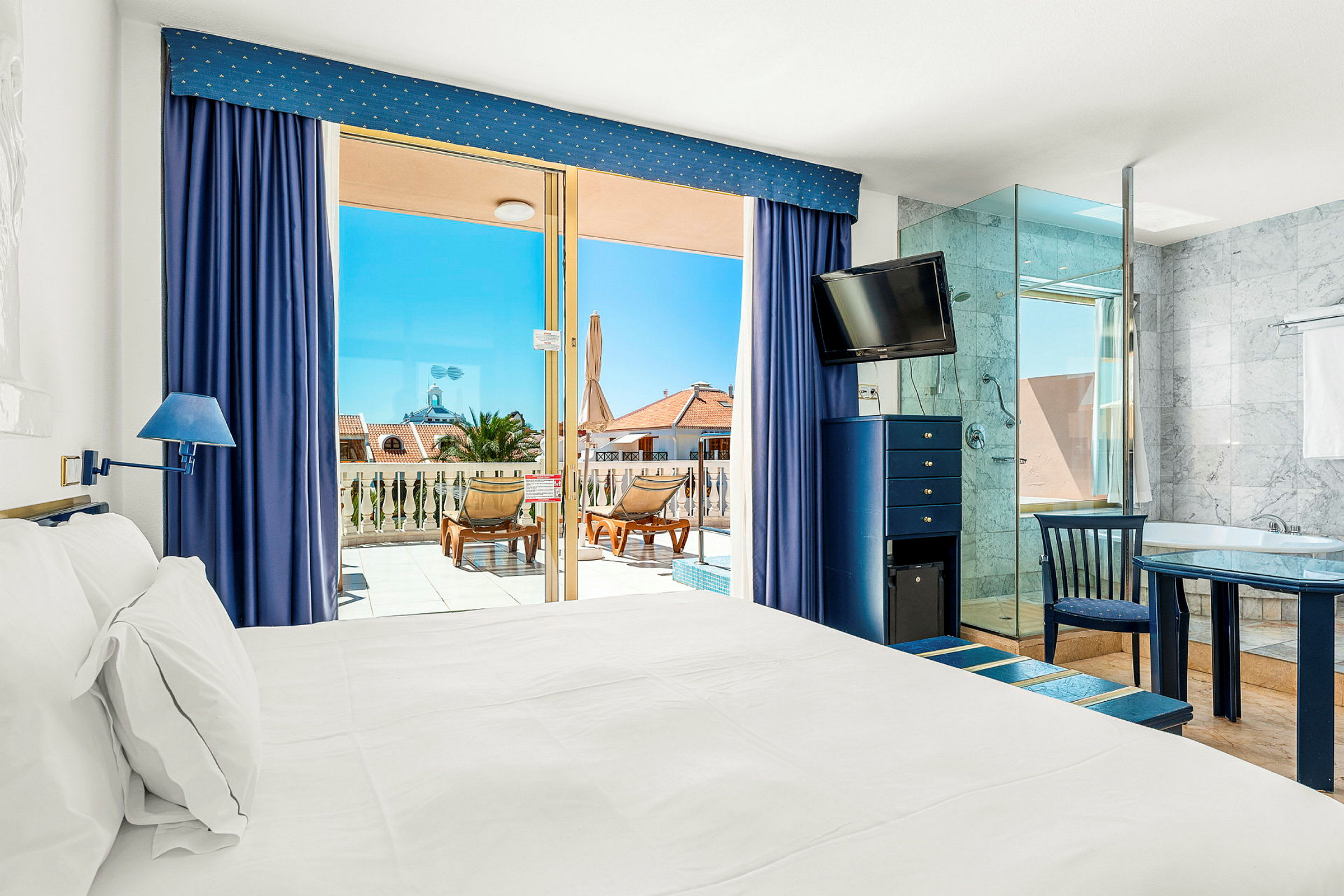 Ett hotellrum med vit säng, blå detaljer och glasdörrar som leder ut till en terrass med solstolar. (AI-genererad text)