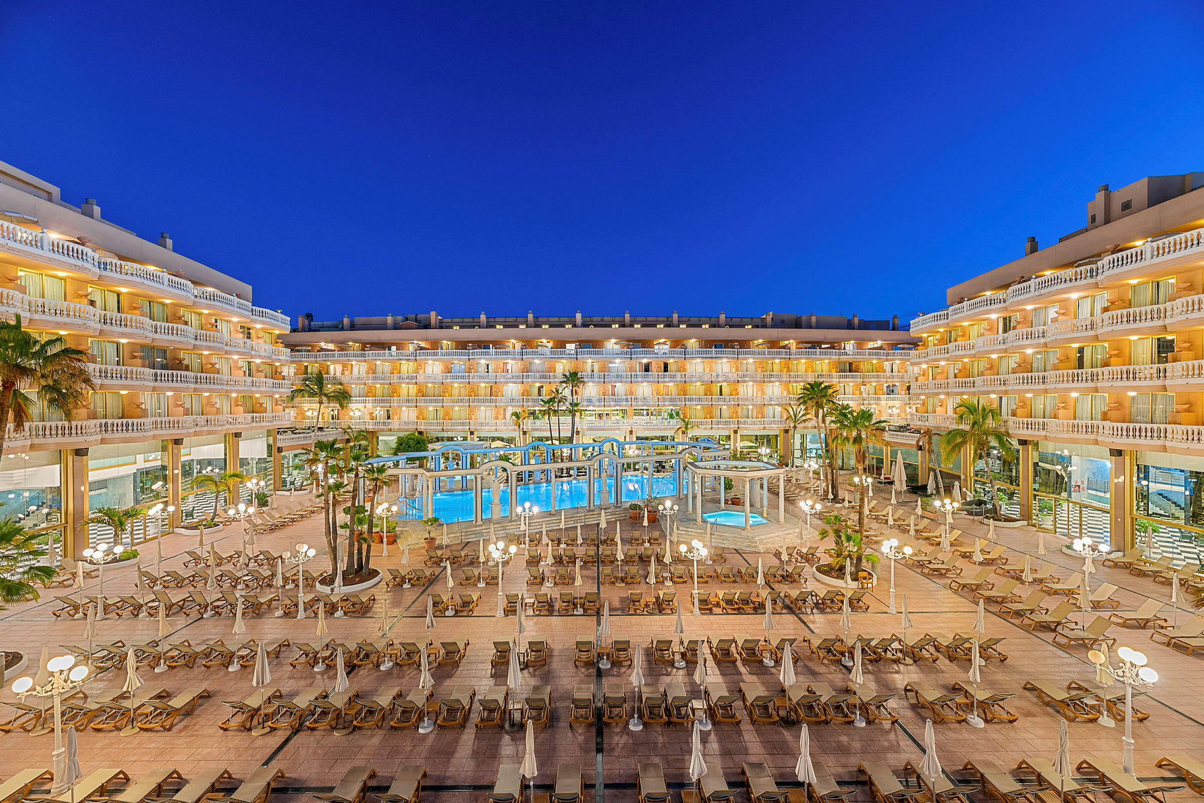 Hotell med upplysta byggnader runt en centralt belägen pool och solstolar. (AI-genererad text)