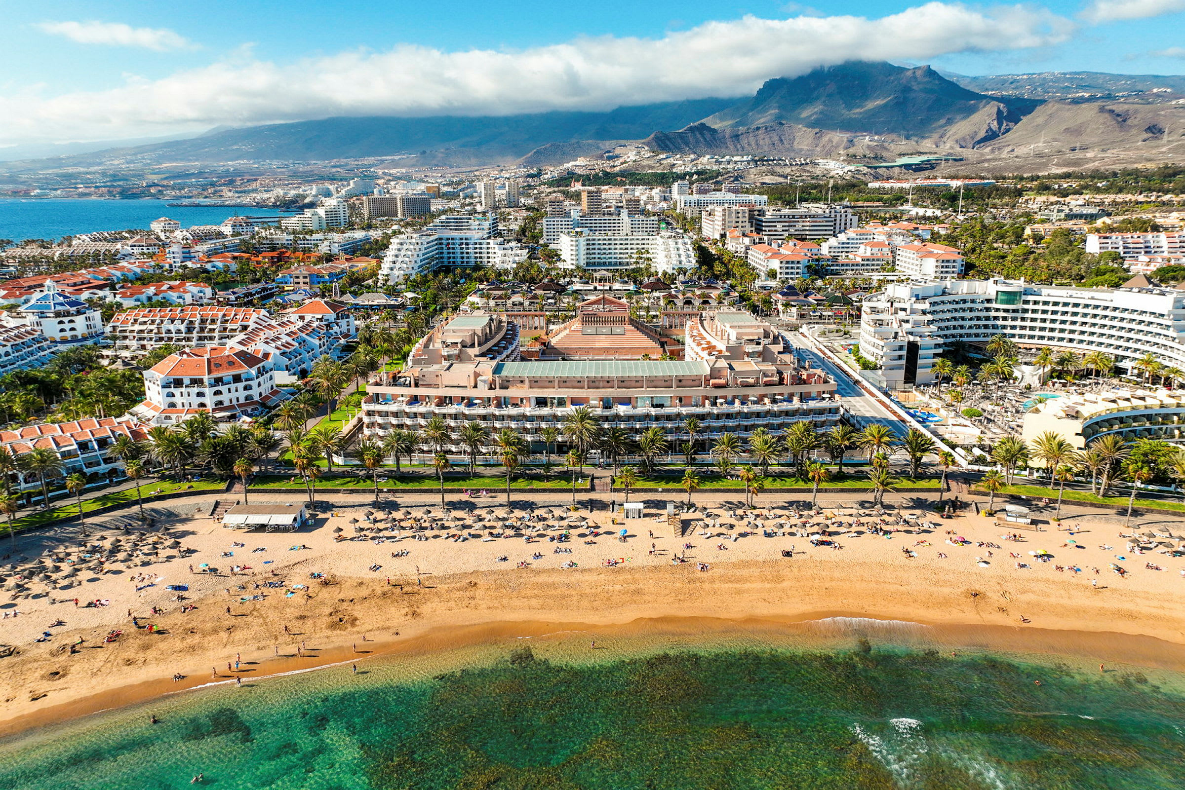 Flygfoto av en strand med solstolar och människor, omgiven av hotell och grönska med berg i bakgrunden. (AI-genererad text)