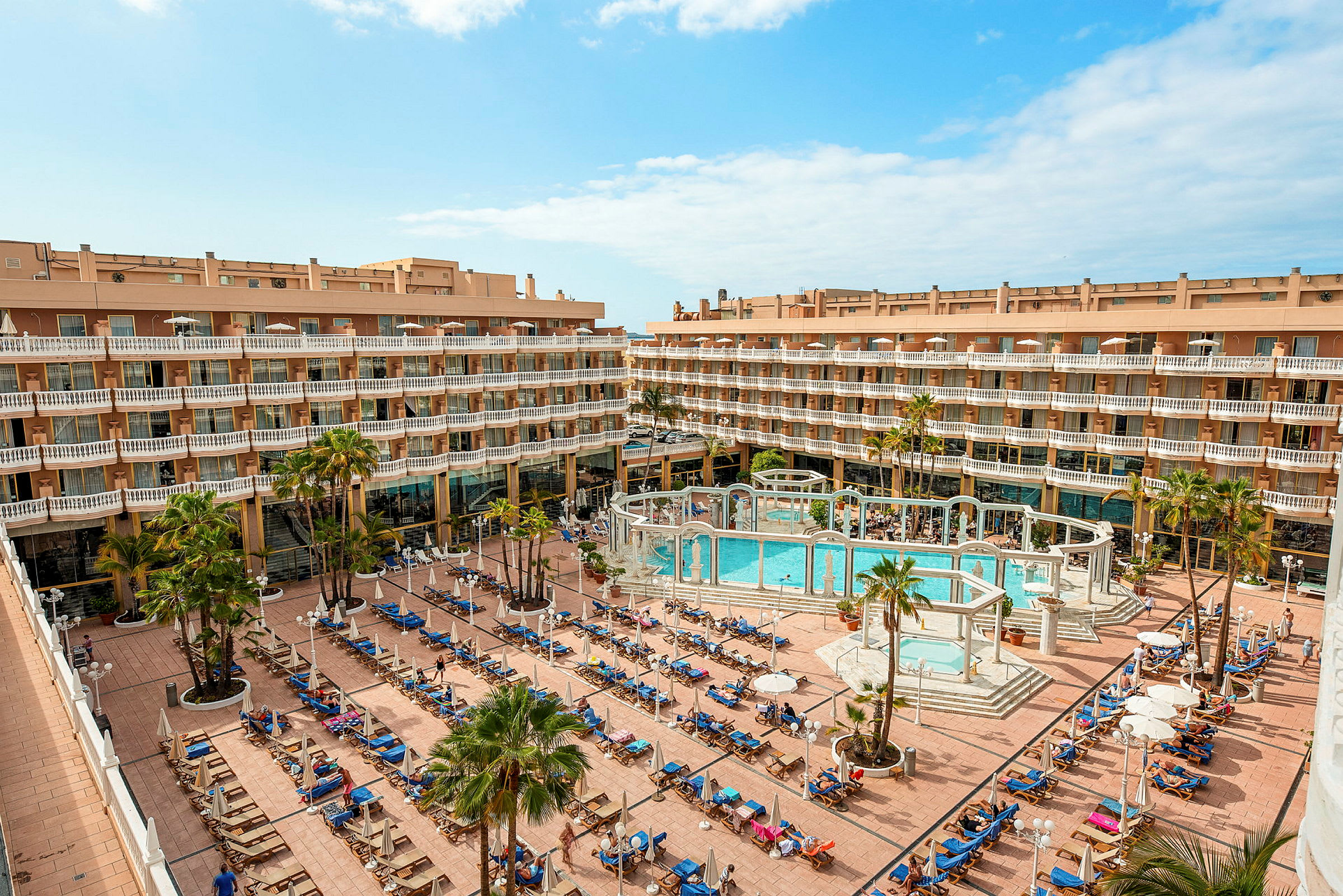 En bild av ett hotellkomplex med en centralt placerad utomhuspool omgiven av solstolar och parasoller. (AI-genererad text)