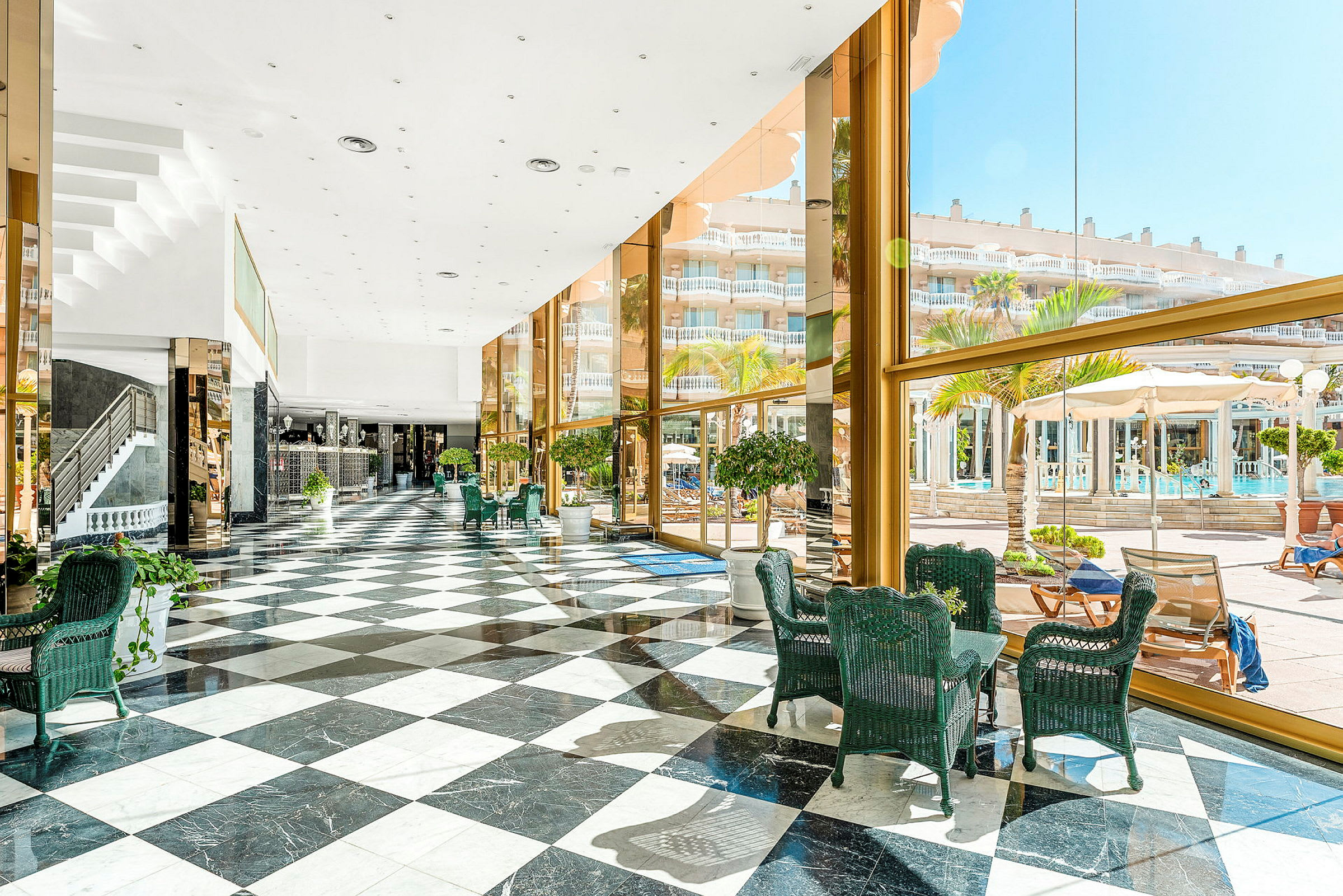 Ett ljust hotellatrium med svartvitt rutigt golv, glasväggar och utsikt över en utomhuspool och palmer. (AI-genererad text)