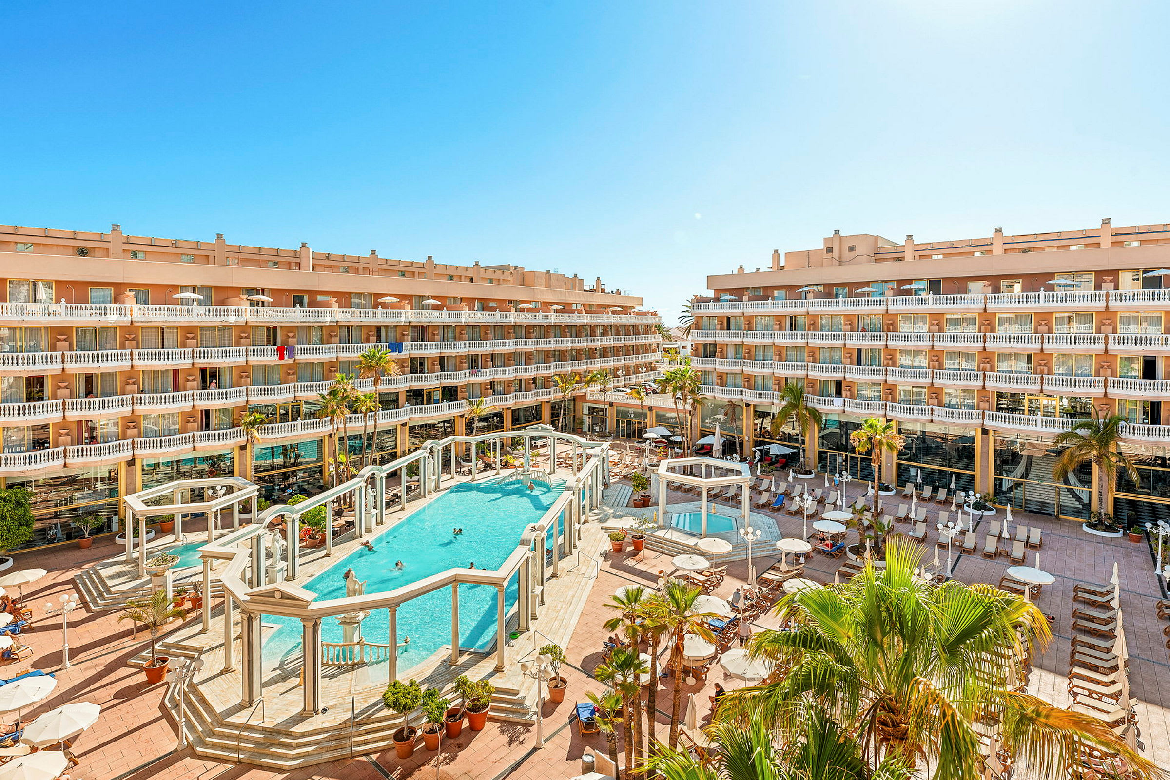 Bild av ett hotellområde med en stor pool i centrum omgivet av byggnader och palmer. (AI-genererad text)