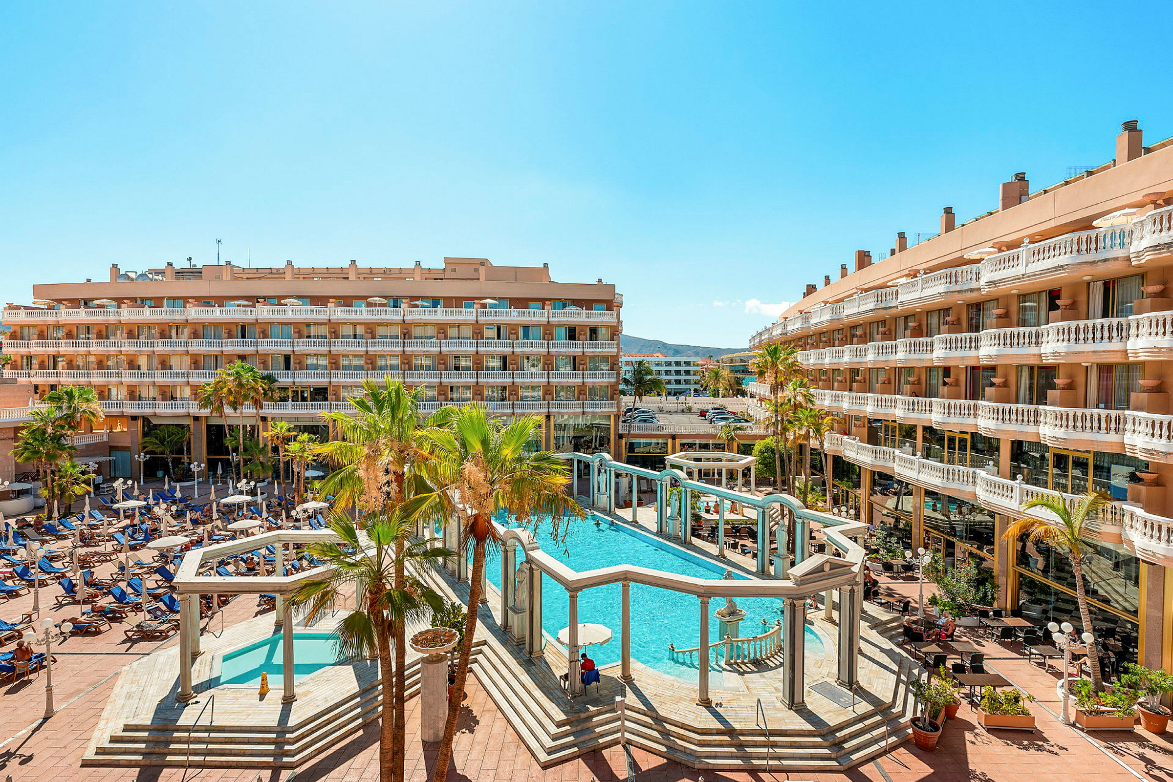 Ett hotellområde med en stor pool omgiven av solstolar och palmer. (AI-genererad text)