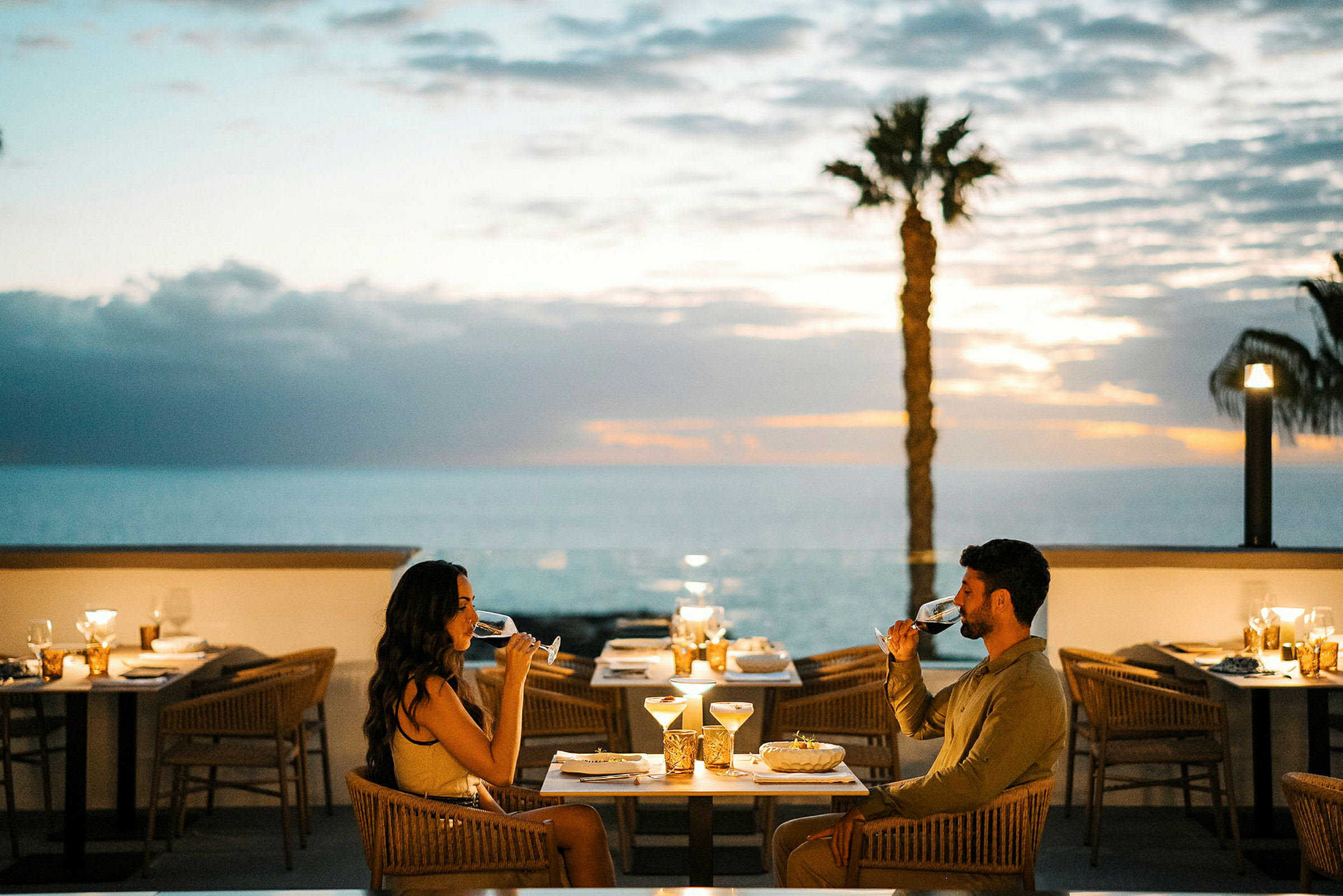 Två personer sitter vid ett dukat bord på en utomhusrestaurang och njuter av ett glas med dryck. I bakgrunden syns havet och en solnedgång. (AI-genererad text)