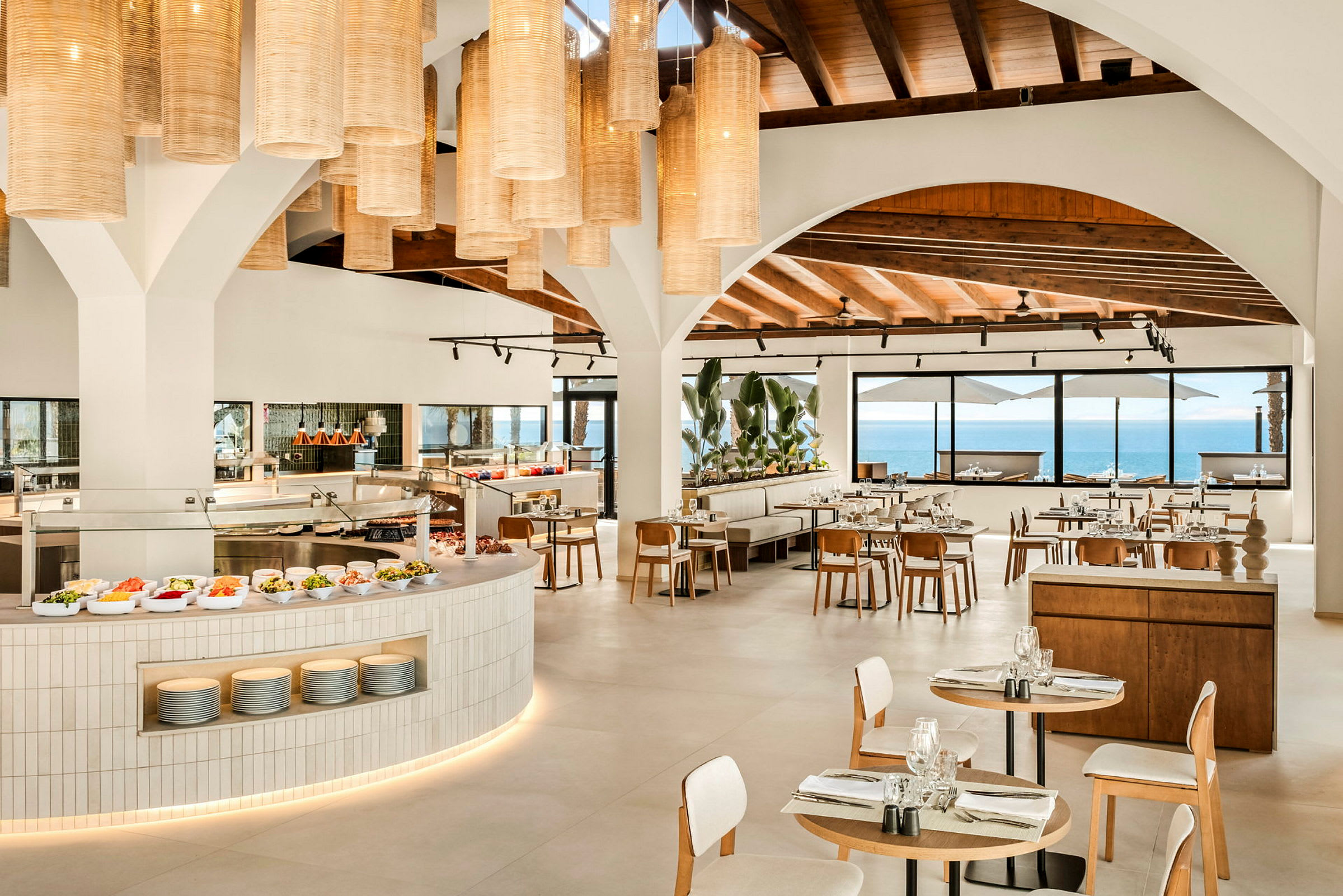 Restaurang med stora bågformade fönster som vetter mot havet, moderna möbler och hängande lampor i taket. (AI-genererad text)