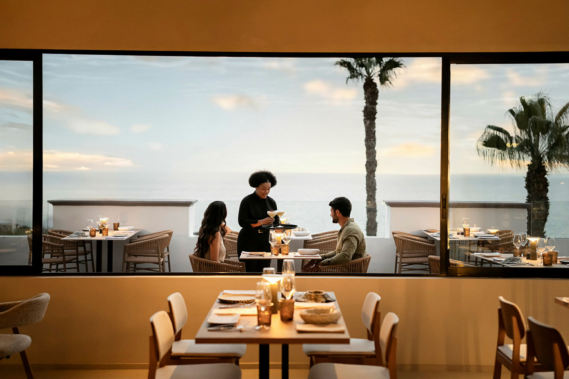 En restaurang med dukade bord, servitör som serverar ett par som sitter vid ett bord. Utsikt över havet och palmer syns genom fönstret. (AI-genererad text)