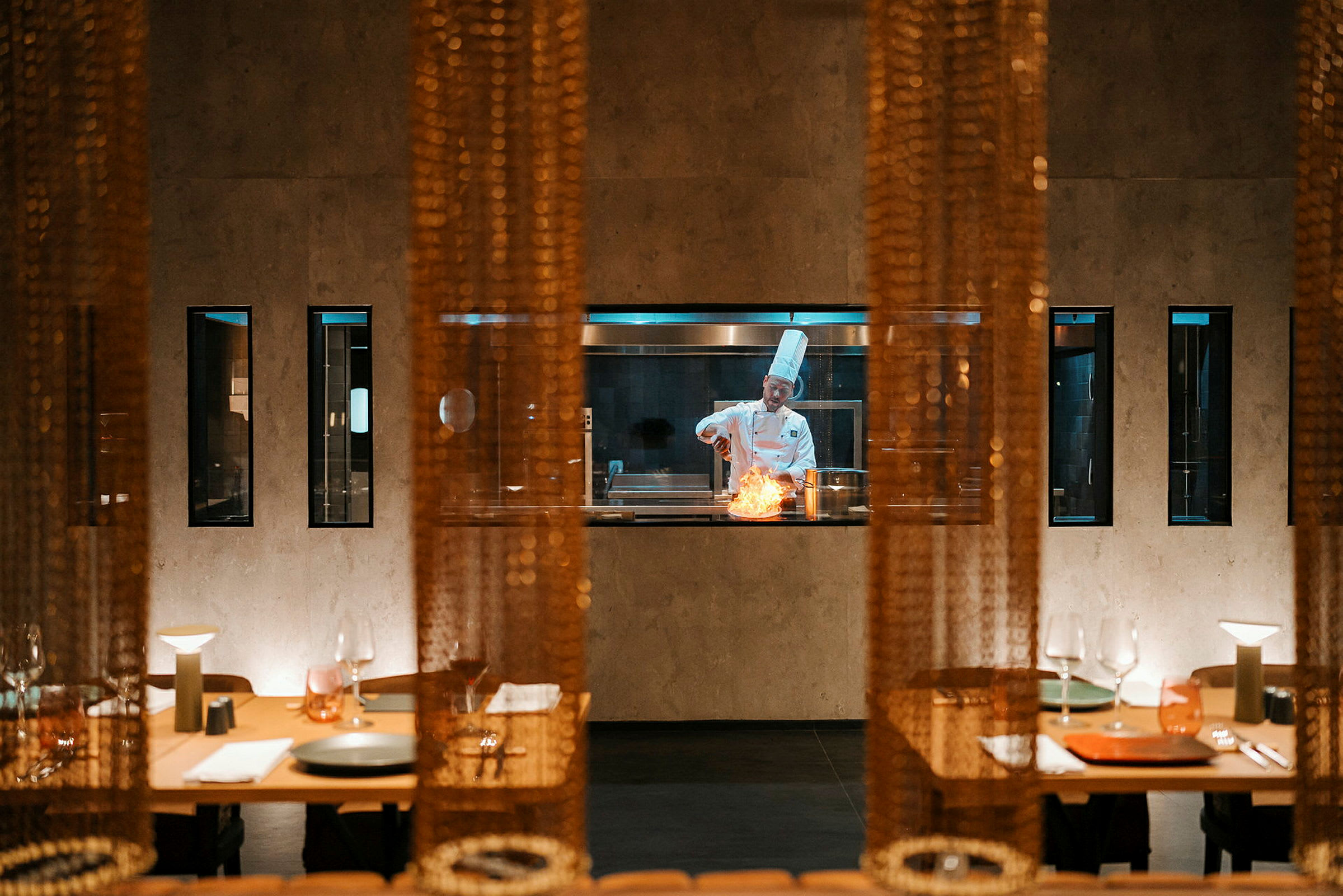 En kock syns innanför en glasvägg i en restaurangmiljö, arbetandes vid en gryta med en flamma. I förgrunden står dukade bord. (AI-genererad text)
