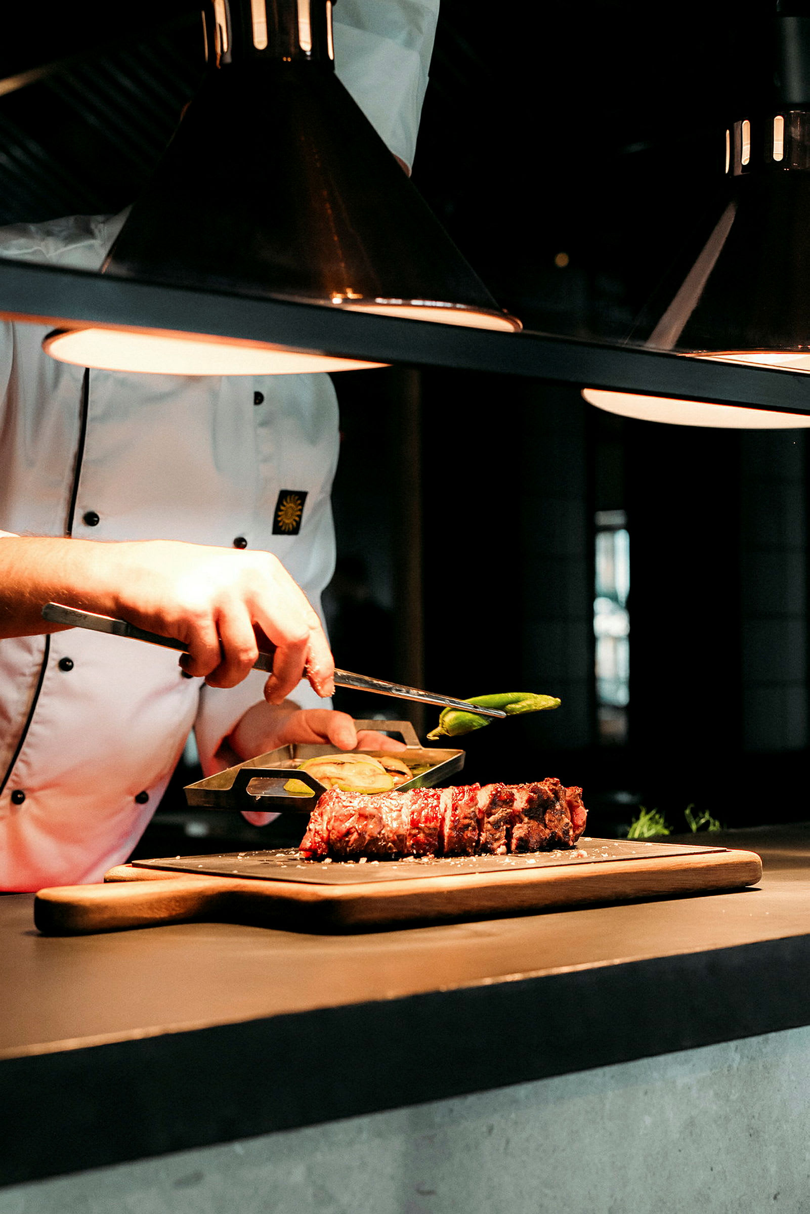 En kock presenterar en maträtt med kött upplagt på en trätallrik, under en arbetslampa. (AI-genererad text)