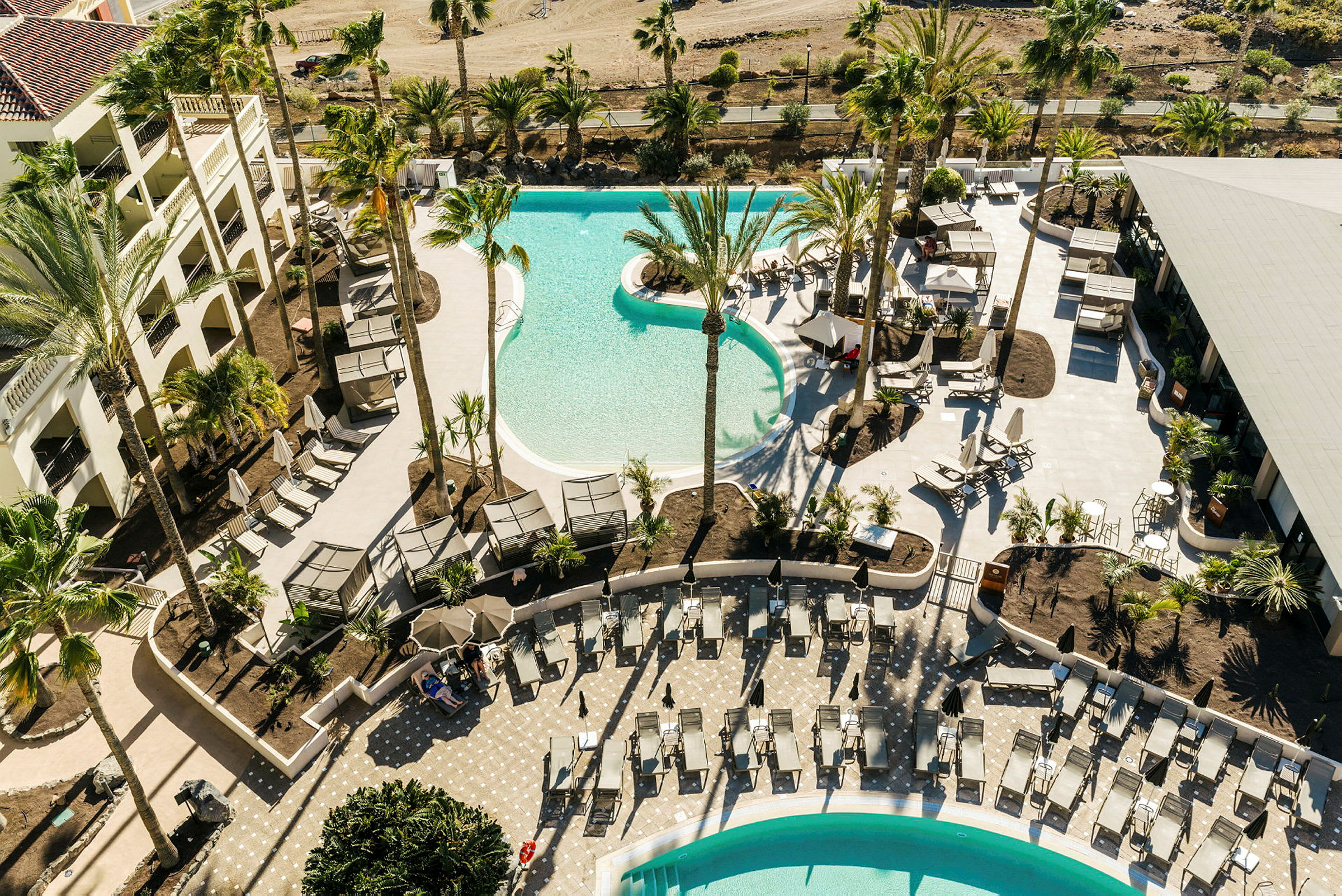 Flygfoto över ett hotellområde med pooler och solstolar omgivna av palmer. (AI-genererad text)