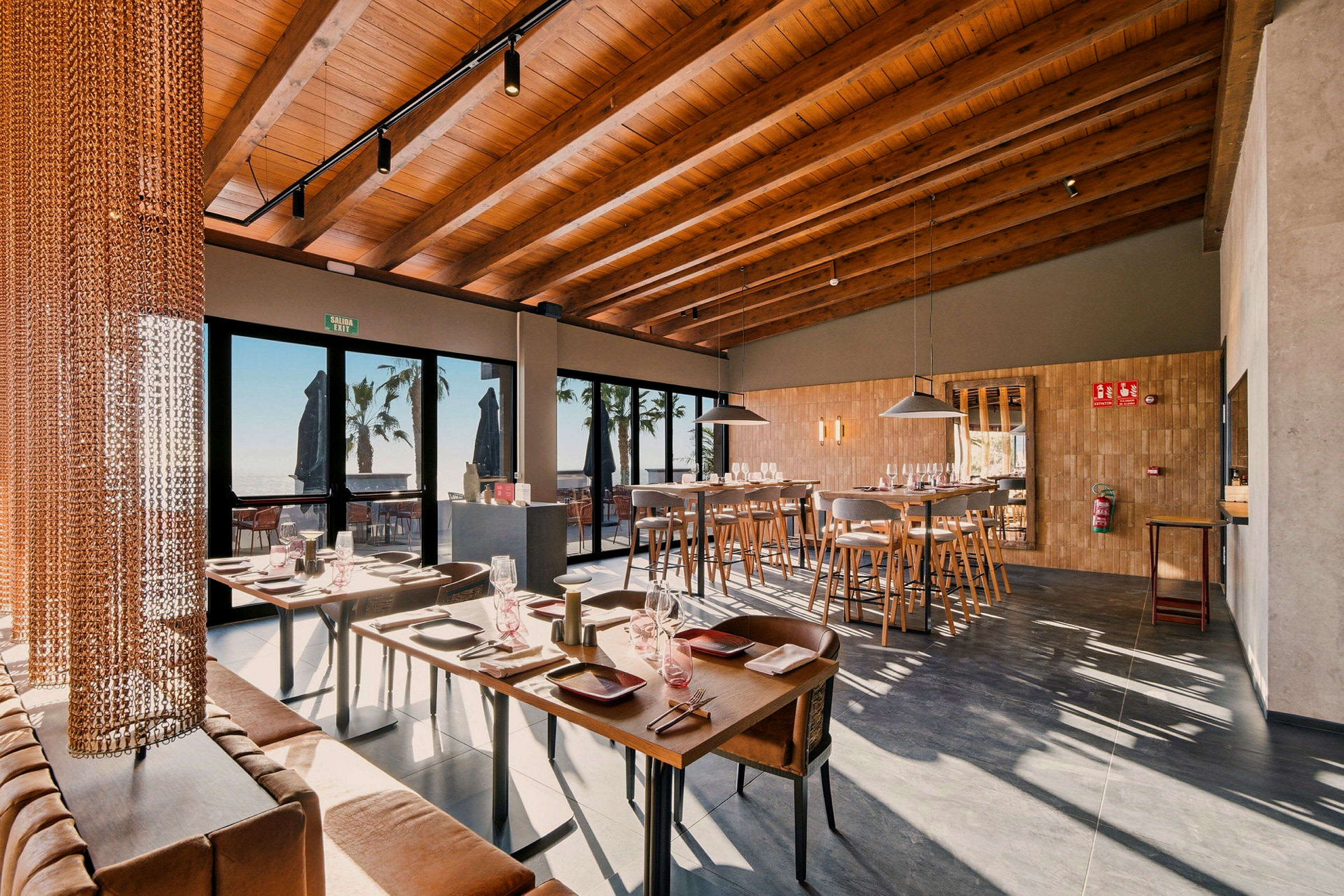 Interiör av en modern restaurang med trämöbler och dukade bord, stora fönster med utsikt utåt. (AI-genererad text)