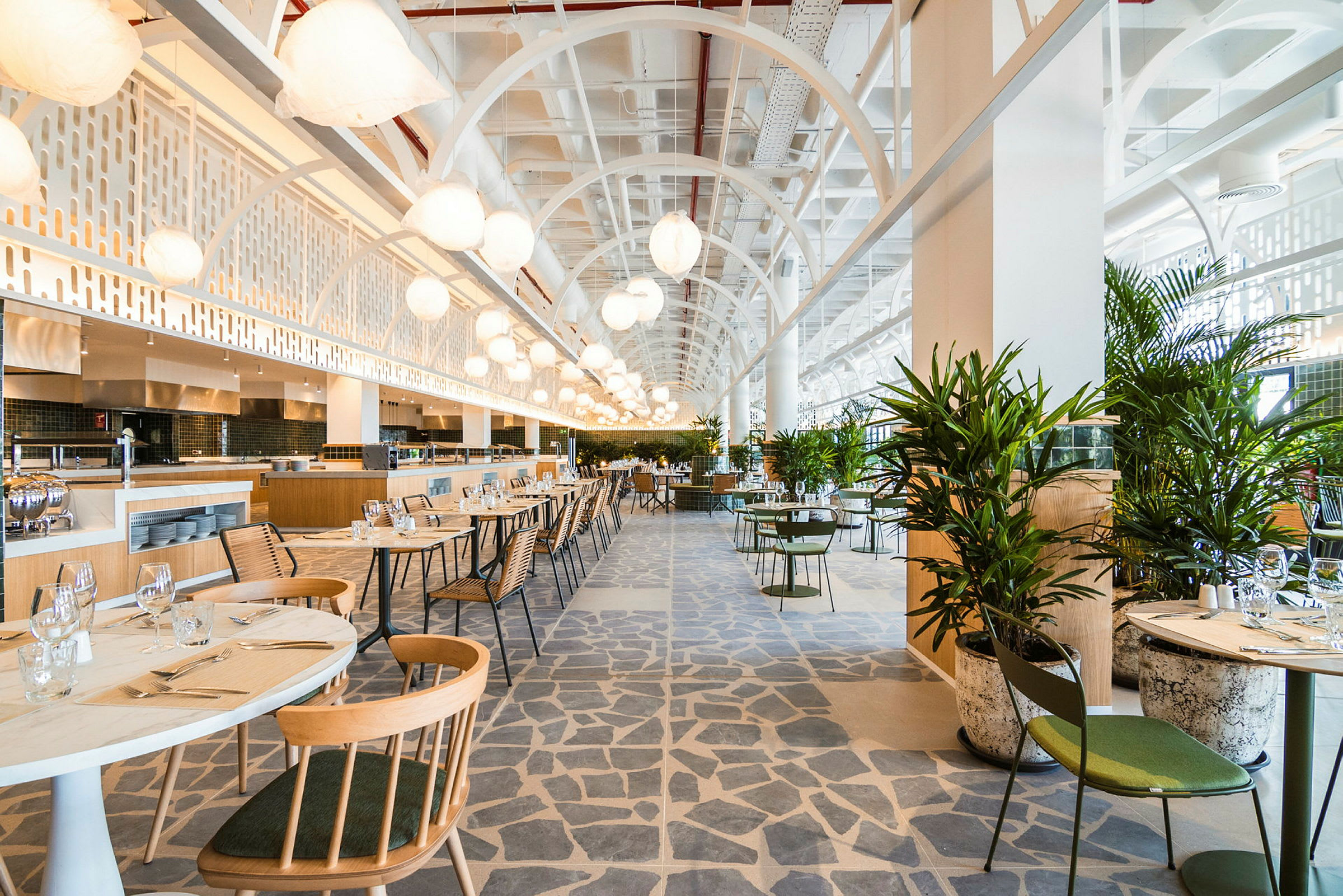 En modern och ljus restaurang med bord och stolar samt gröna växter. Takhöjd och belysning bidrar till en luftig atmosfär. (AI-genererad text)