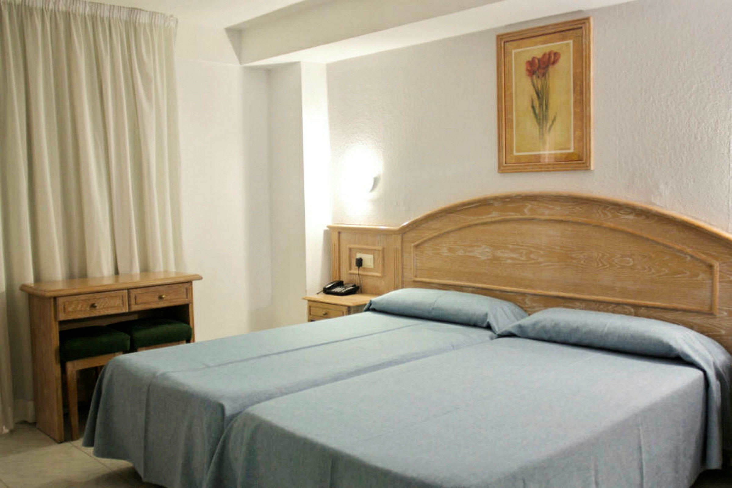 Hotellrum med två enkelsängar med blåa sängkläder. Trämöbler och en tavla pryder väggen. (AI-genererad text)