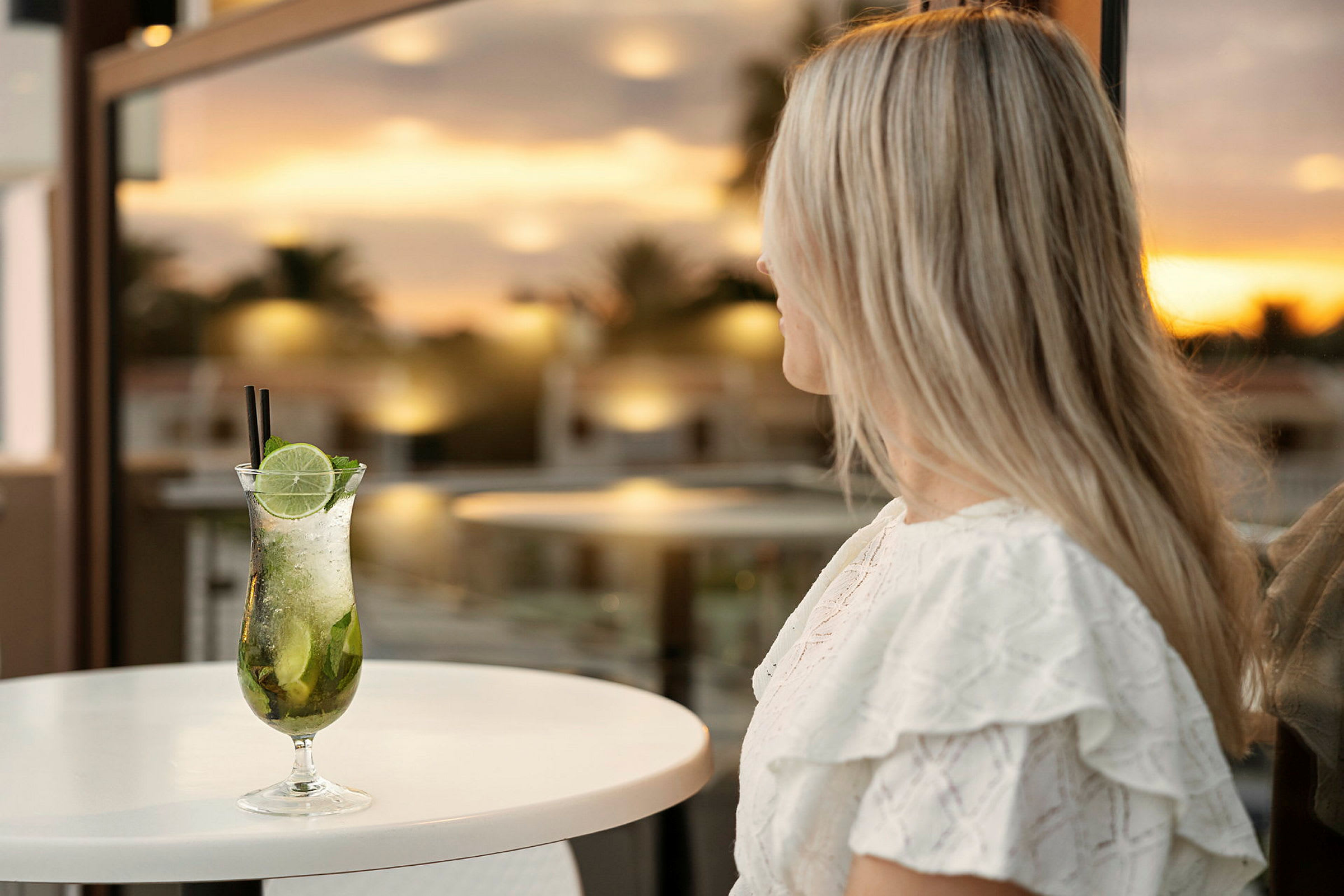 Kvinna med långt ljust hår sitter vid ett bord med en cocktail framför sig, bakgrund är en solnedgång. (AI-genererad text)