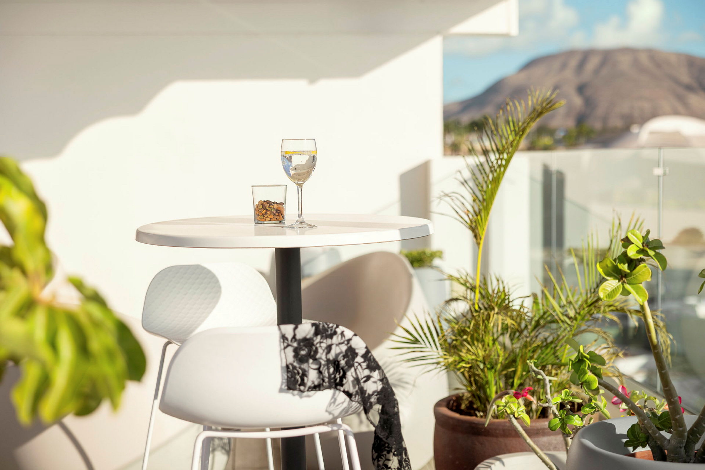 Ett litet bord på en terrass, med ett glas dryck och en skål nötter i solen, omgiven av växter och en utsikt över ett berg. (AI-genererad text)