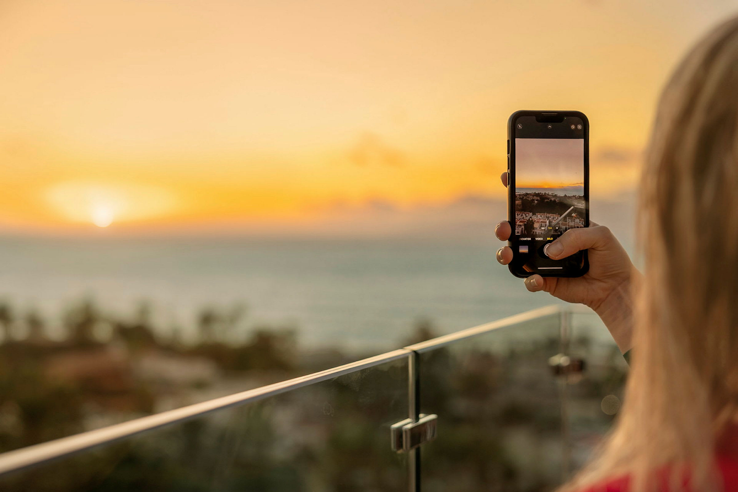 Person tar en bild med sin mobilkamera av en solnedgång över havet från en balkong. (AI-genererad text)