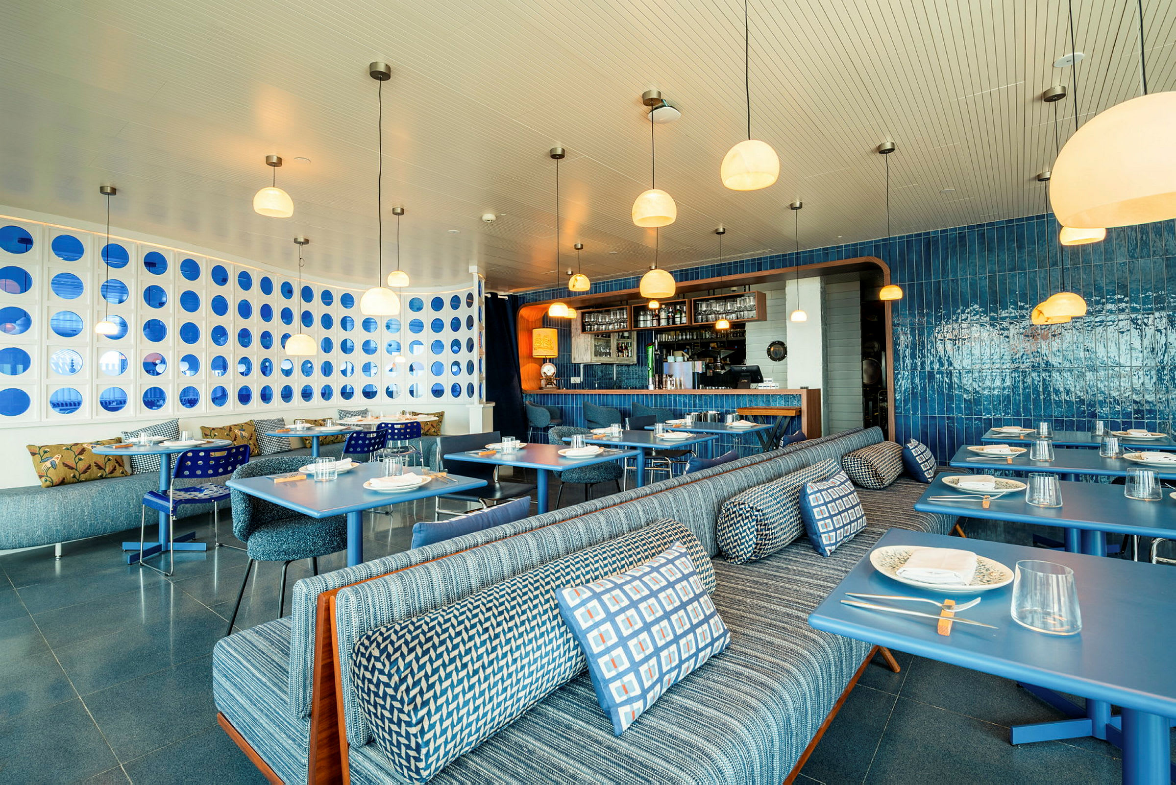 Bild av en modern restaurang med elegant inredning i blå toner, sittplatser med mönstrade dynor och dukade bord. (AI-genererad text)