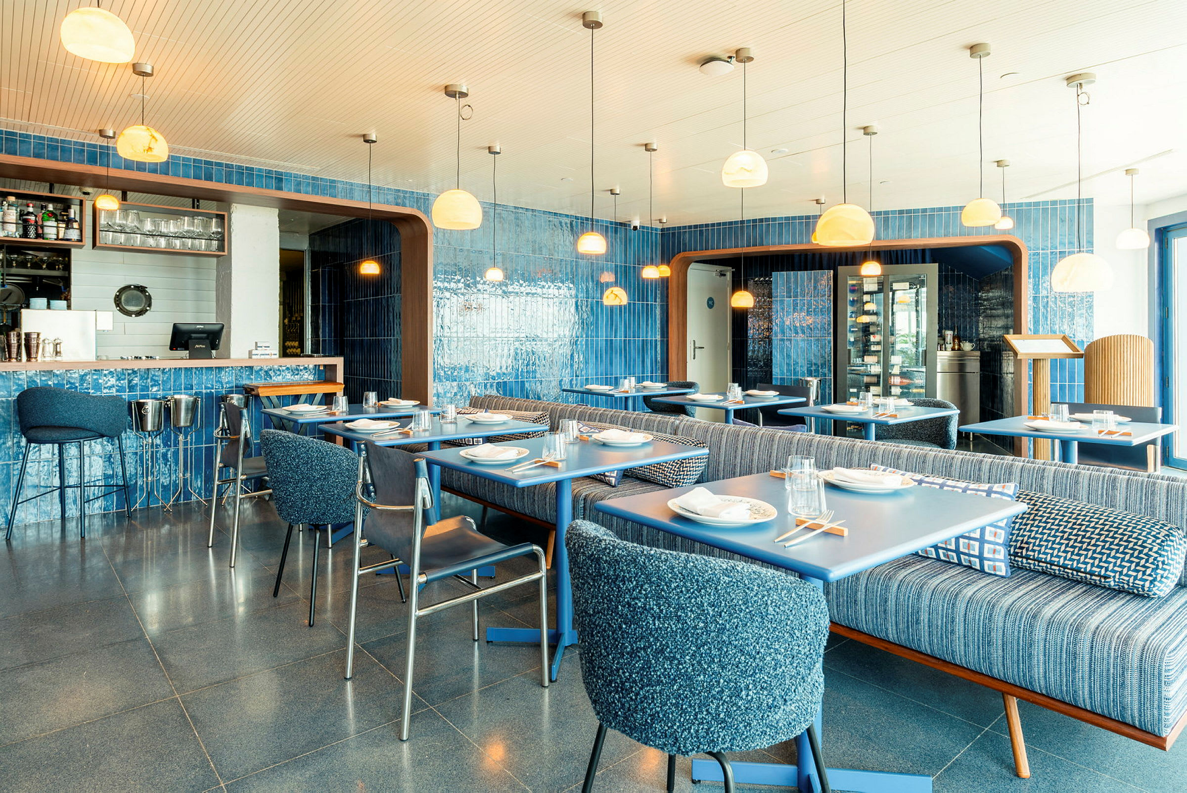 En färgglad restauranginteriör med blå väggar, bord och stolar. Ljusa lampor hänger från taket. (AI-genererad text)
