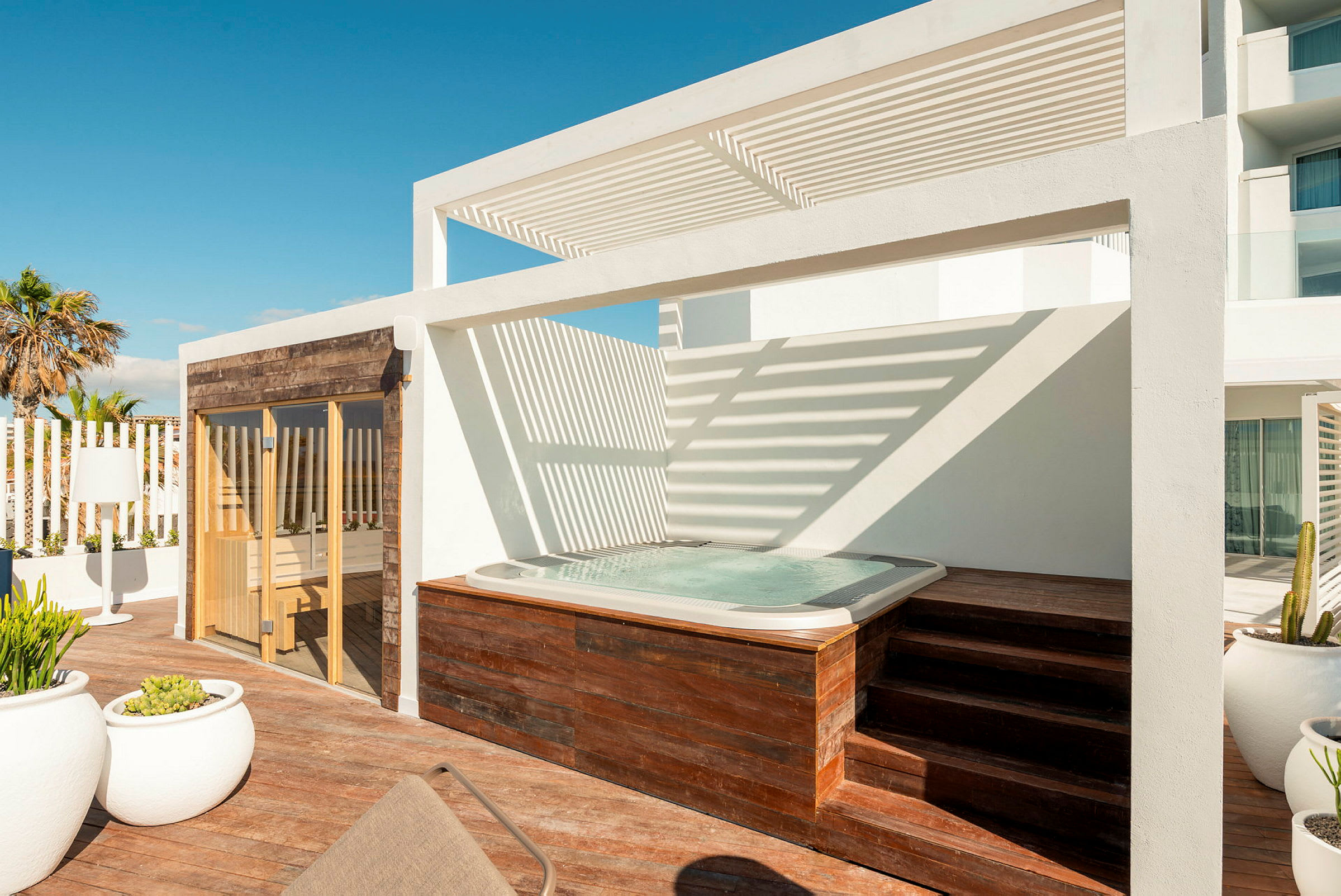 En terrass med en bubbelpool, en vit pergola, och en bastu med träbeklädd fasad. (AI-genererad text)