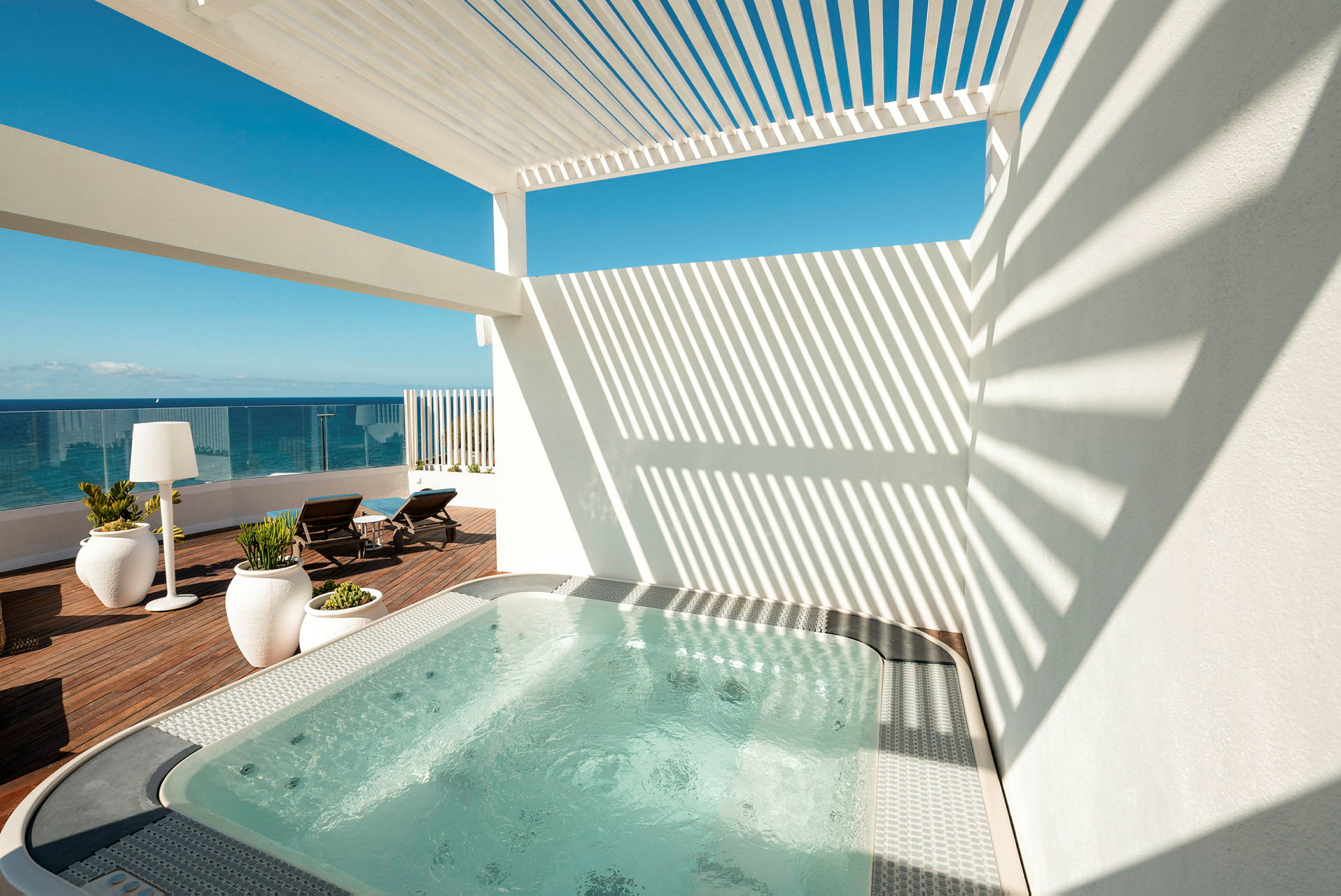 En terrass med bubbelpool och utsikt över havet. Terrassen har solstolar, stora växter i krukor och en pergola som ger skuggiga områden. (AI-genererad text)