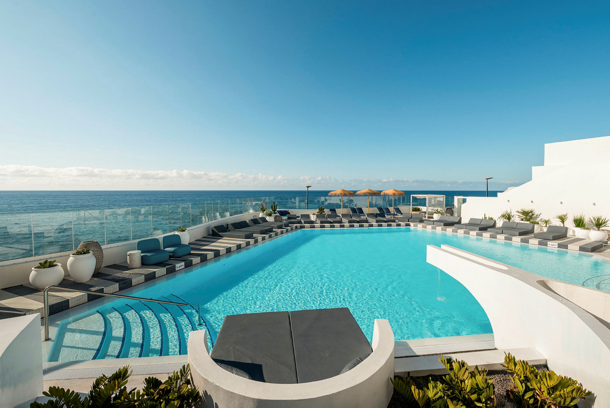 En pool med turkost vatten omgivet av solstolar och utsikt över havet. I bakgrunden syns en klar blå himmel. (AI-genererad text)