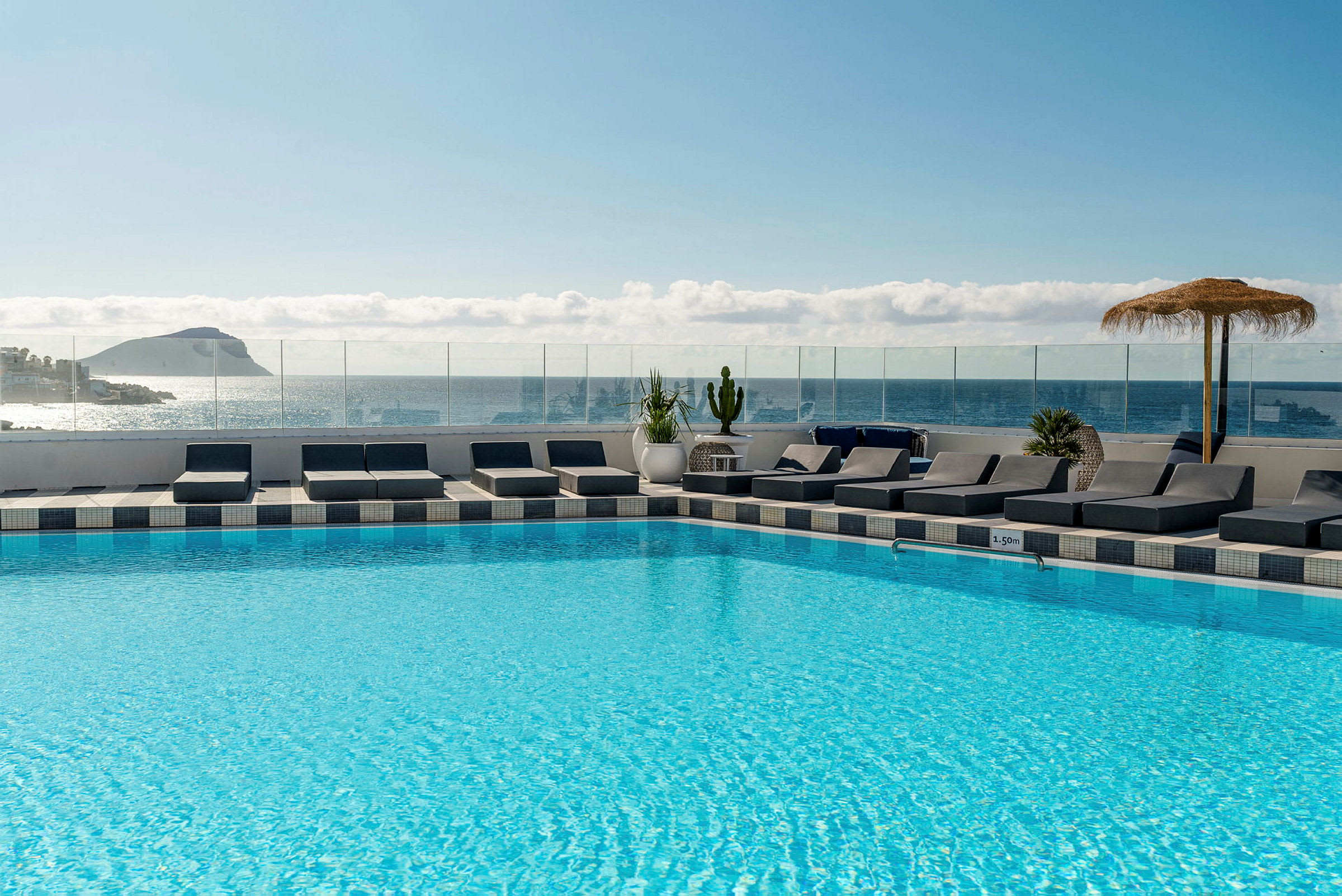 En pool på ett hotell med solstolar runtom samt utsikt över havet och en ö i bakgrunden. (AI-genererad text)